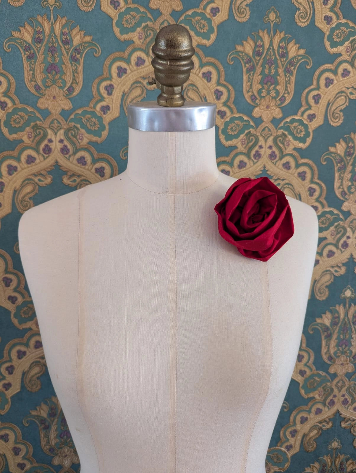 Broochella Isabella Rossellini handmade silk rosette flower brooch in red. Pictured on a mannequin.  The background is green and gold damask wallpaper