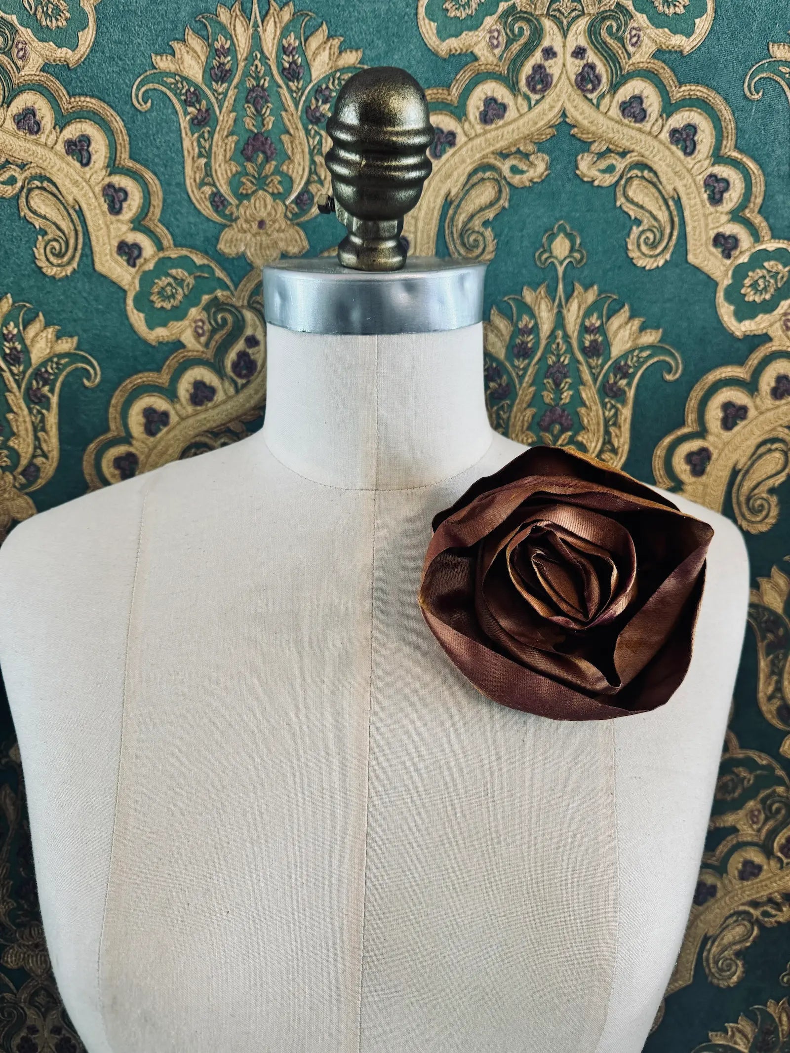 Broochella Isabella Rossellini handmade silk rosette flower brooch in rust. Pictured on a mannequin.  The background is green and gold damask wallpaper