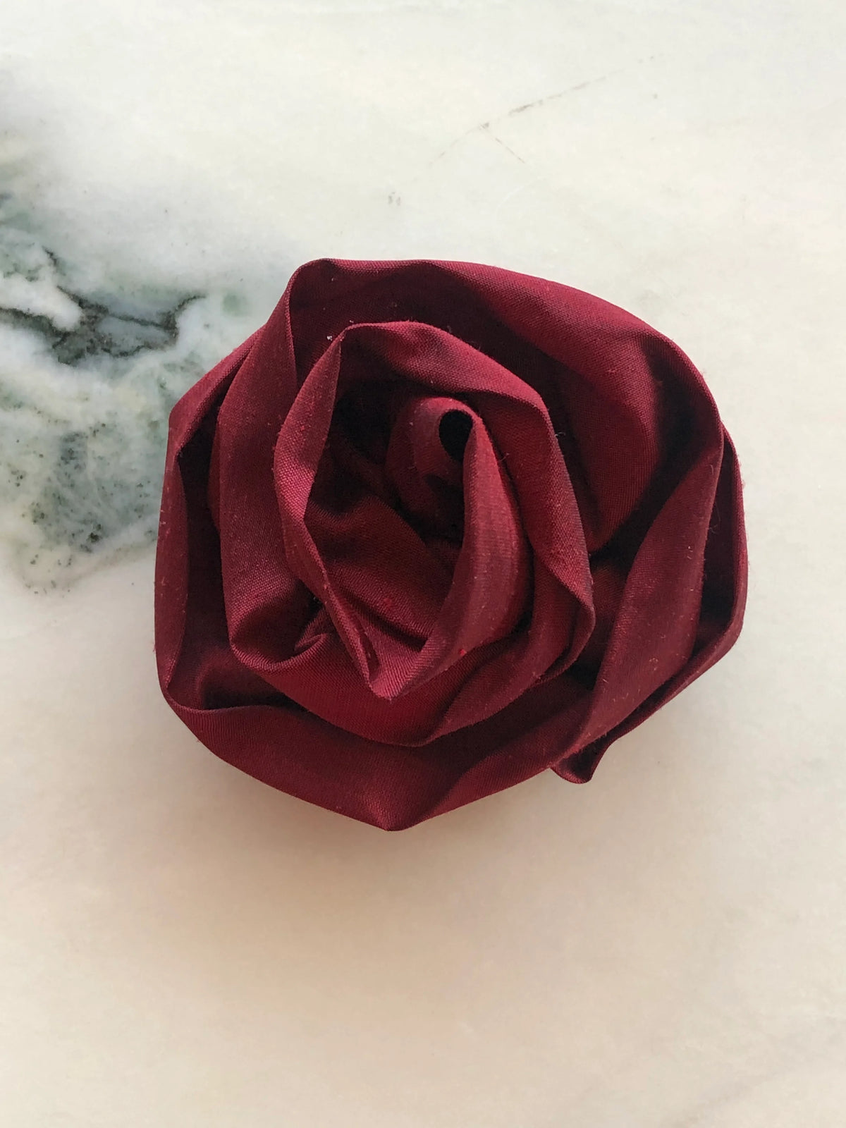 Handmade Broochella Isabella Rossellini designer silk rosette brooch in deep burgundy, sculpted into layered rose petals and photographed on white marble.