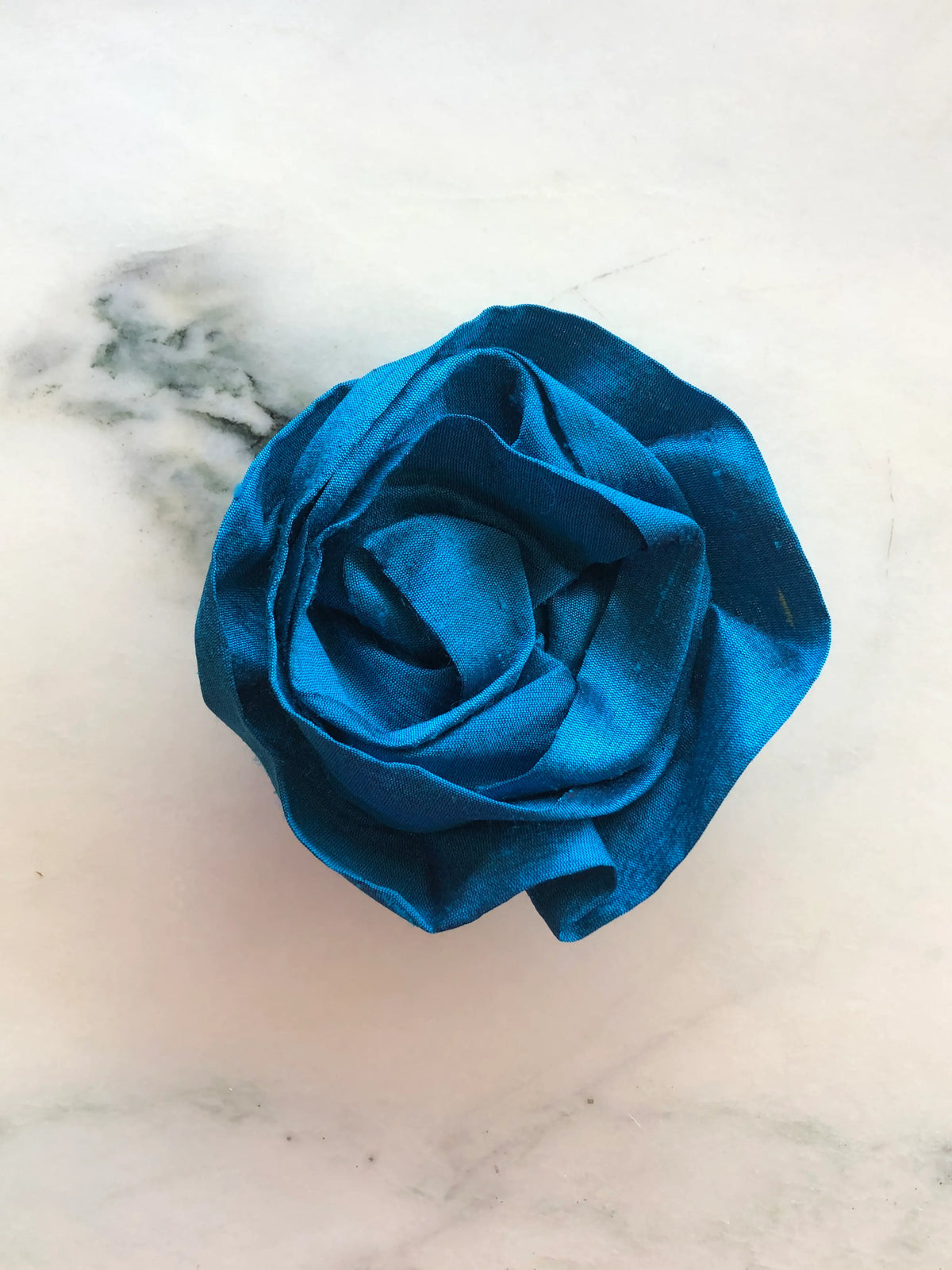 Handmade Broochella Isabella Rossellini designer silk rosette brooch for men in peacock blue, featuring sculptural folded petals for statement lapel styling, photographed on white marble.