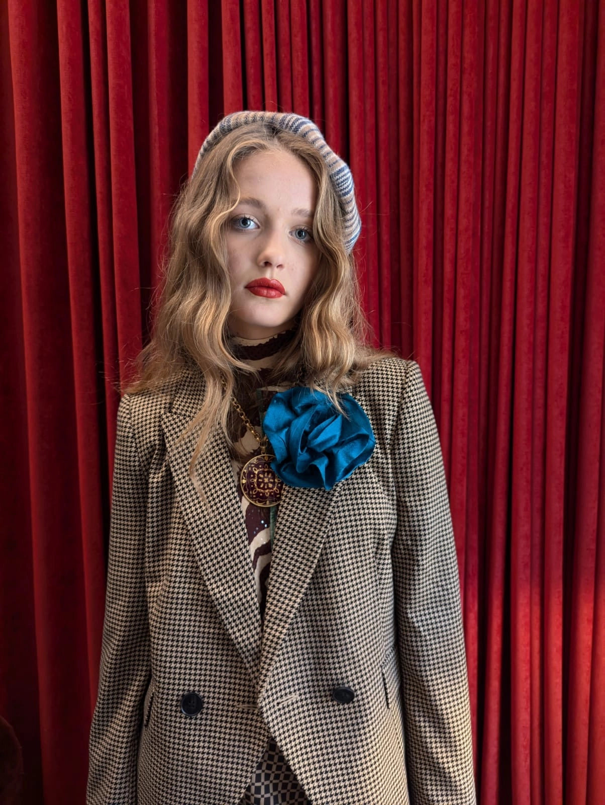 A young woman stands in front of a red velvet curtain wearing a beige and black houndstooth blazer, a Broochella peacock blue isabella rossellini fabric flower broochaccessory, a striped beret, and a patterned top.