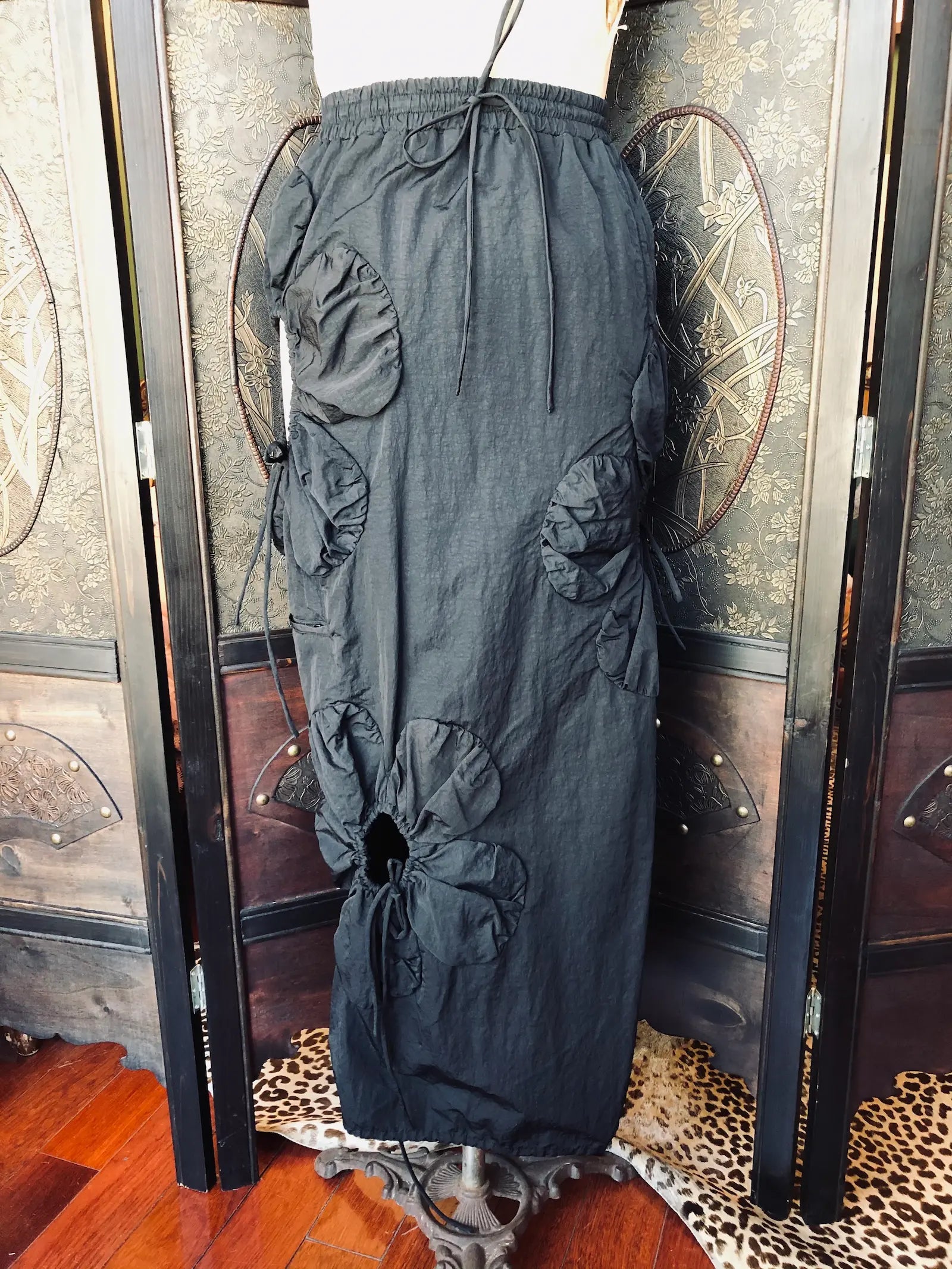 Broochella J Kim Small, 100% nylon, Black
Elasticated drawstring waist. Sculptural rosette appliqué detailing
Adjustable drawstring accents
Lightweight non stretch fabric. It is presented on a vintage mannequin. The background is a vintage, decorative, wooden room divider. 