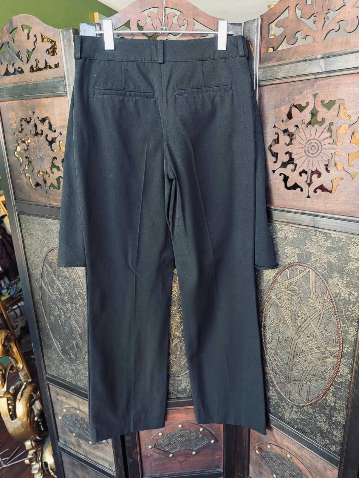 Broochella J.W. Anderson black trousers with an attached skirt overlay. They are  hanging on a wooden hanger against a decorative carved wooden screen.