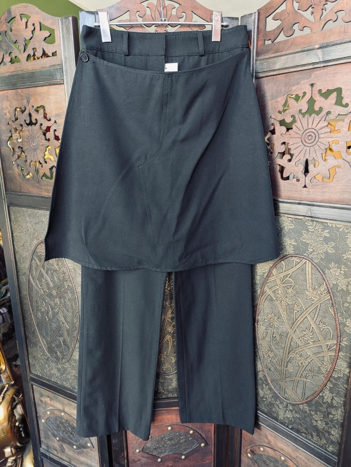 Broochella J.W. Anderson black trousers with an attached skirt overlay. They are  hanging on a wooden hanger against a decorative carved wooden screen.