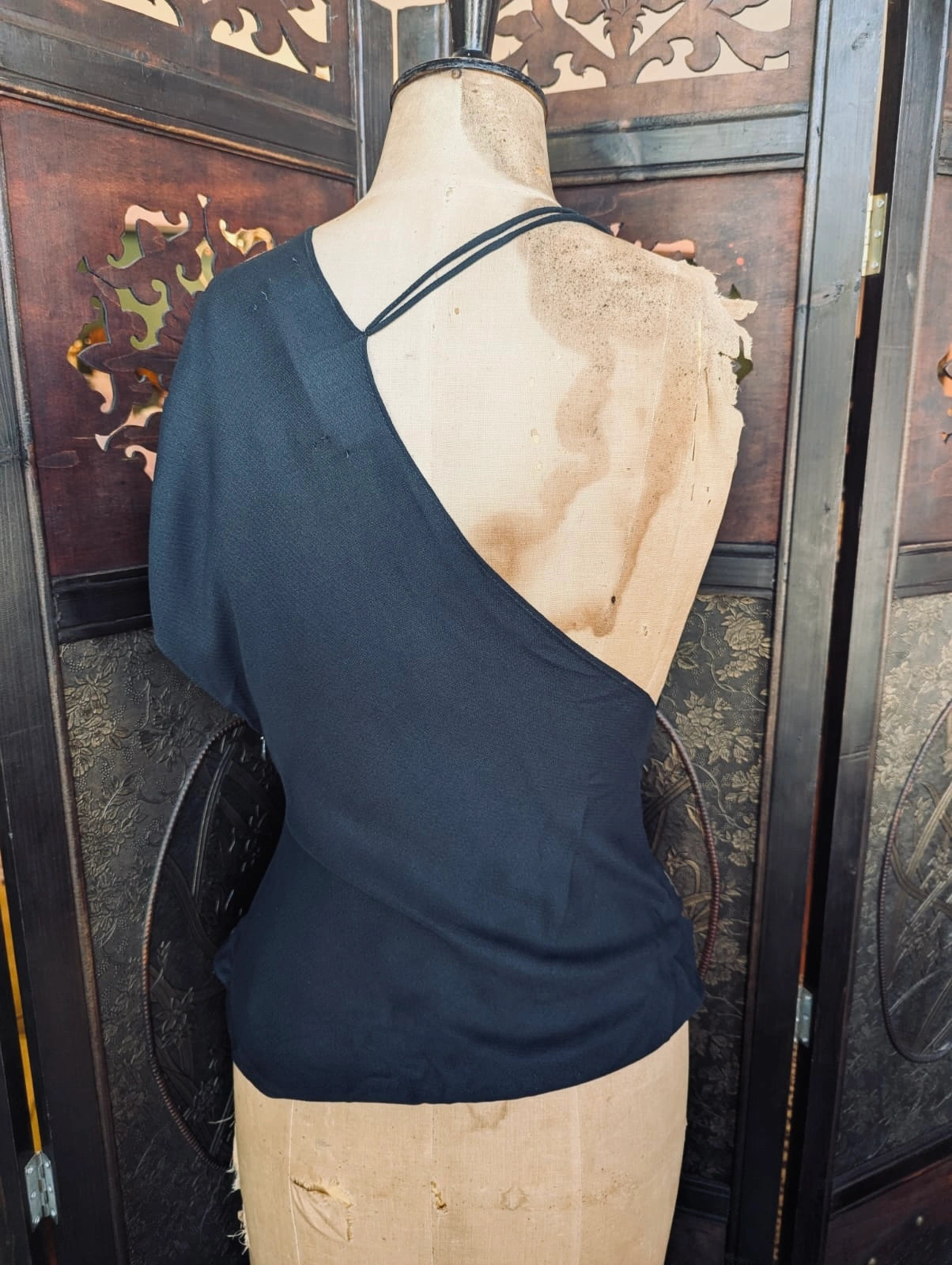 The back view of Broochella designer jacquemus le souk black multi wear draped top. It is pictured on a mannequin.The background is an ornate dark wood decorative room divider. 