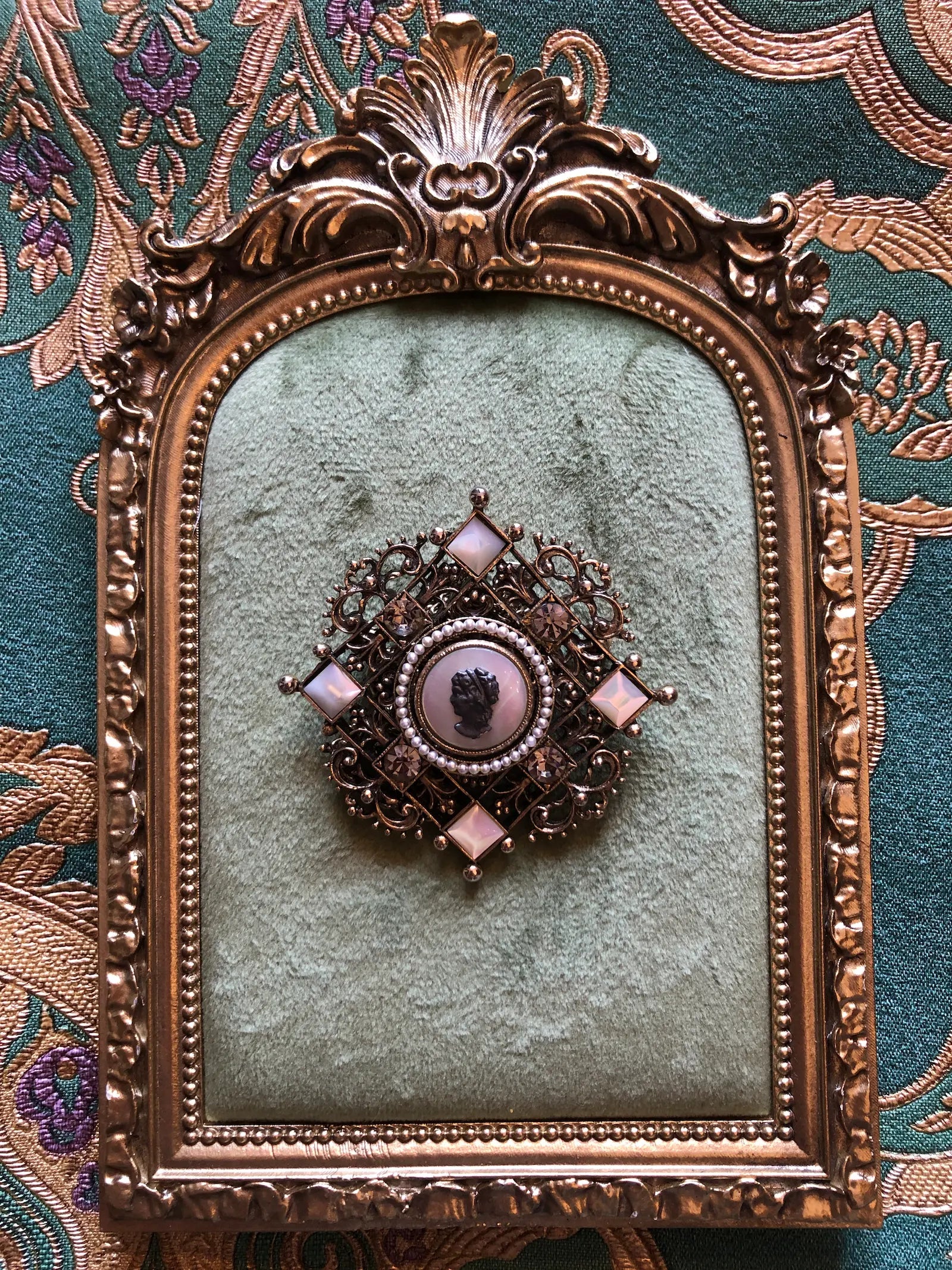 Broochella Kaiserin vintage German cameo brooch pendant necklace with ornate gold-tone filigree, seed pearls and crystal accents.. It is displayed on a green velvet surface inside an antique gold frame against a patterned green textile background.