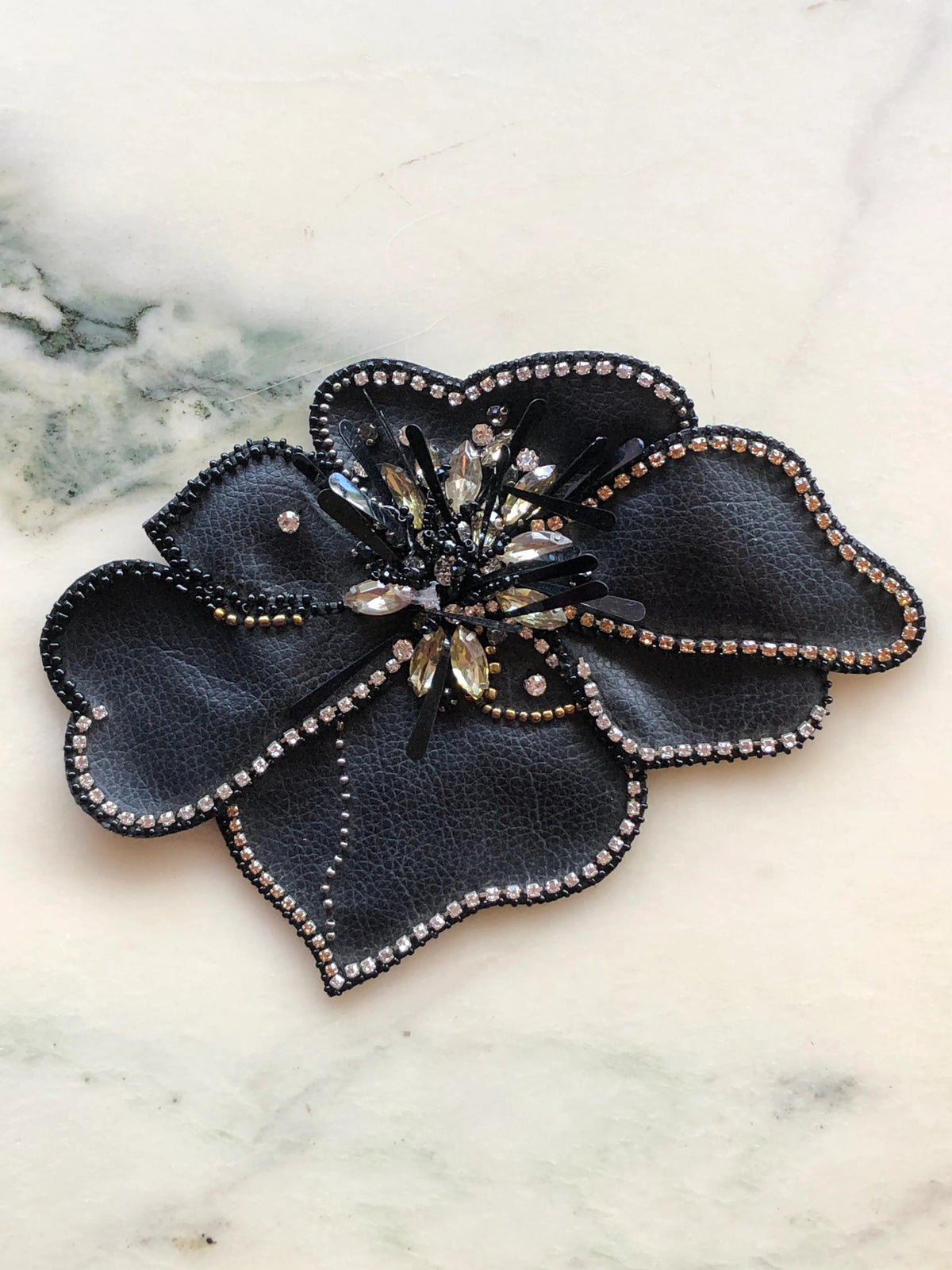 Broochella Koi Bejewelled Flower Brooch Pin in black, featuring sculptural leather-look petals edged with sparkling crystals, beads and a dramatic crystal and sequin centre, photographed on a white marble background with soft grey-green veining.