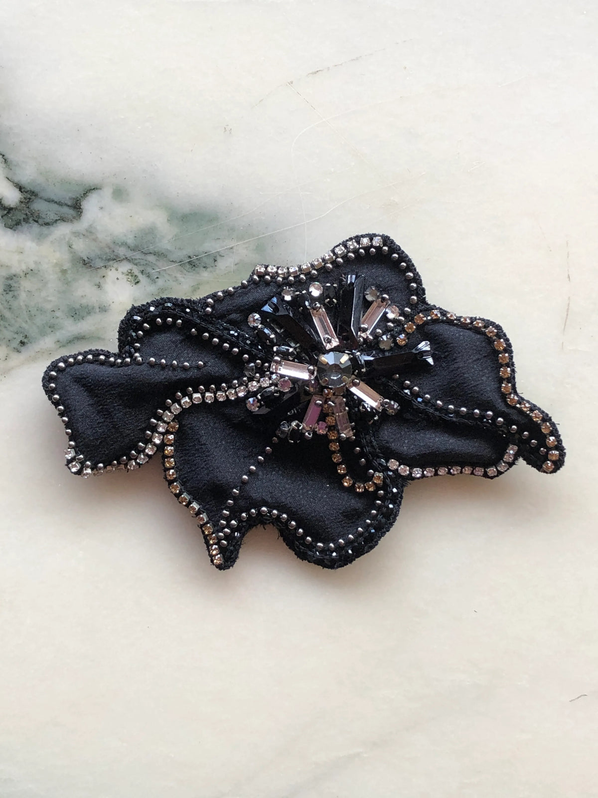 Broochella Koi Bejewelled Flower Brooch Pin in black, featuring softly folded fabric petals embellished with silver beads, crystals and black sequins around a jewelled centre, photographed on a white marble background with soft grey-green veining.
