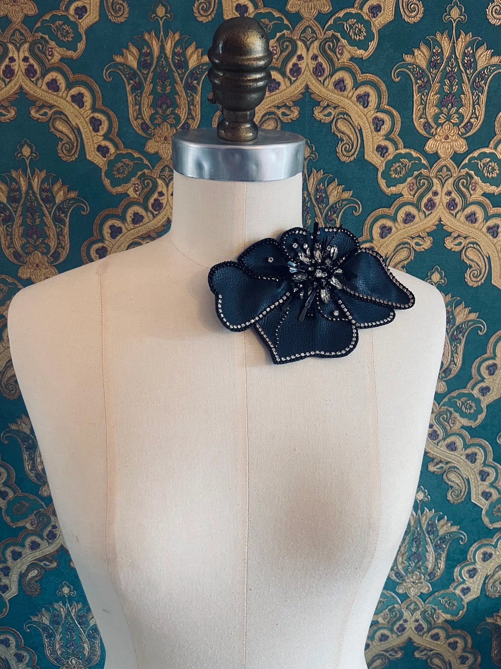 Broochella Koi large black leather flower brooch with hand beaded crystal embellishment. Pictured on a mannequin. The background is green and gold damask wallpaper