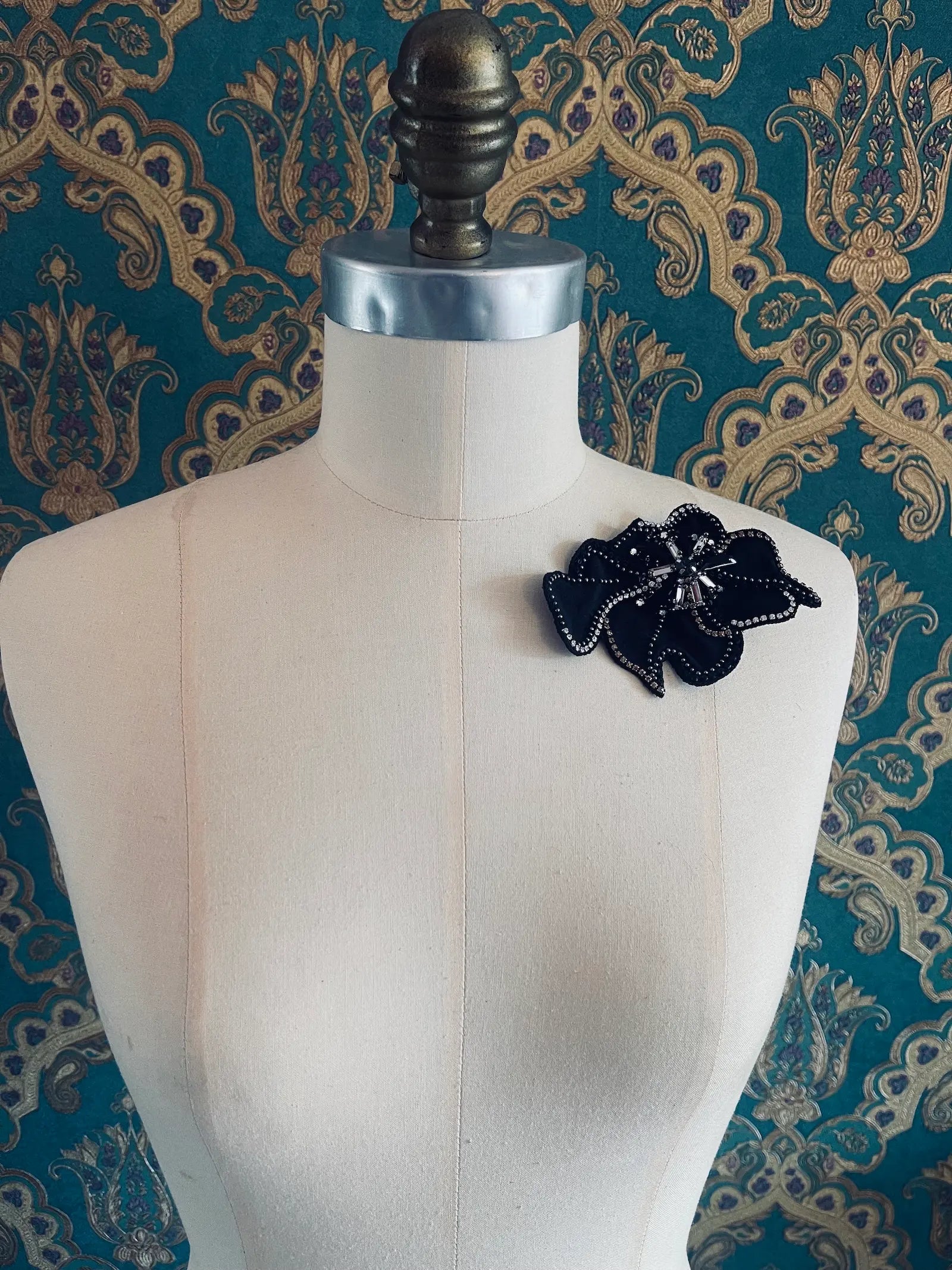 Broochella Koi small black leather flower brooch with hand beaded crystal embellishment. Pictured on a mannequin. The background is green and gold damask wallpaper