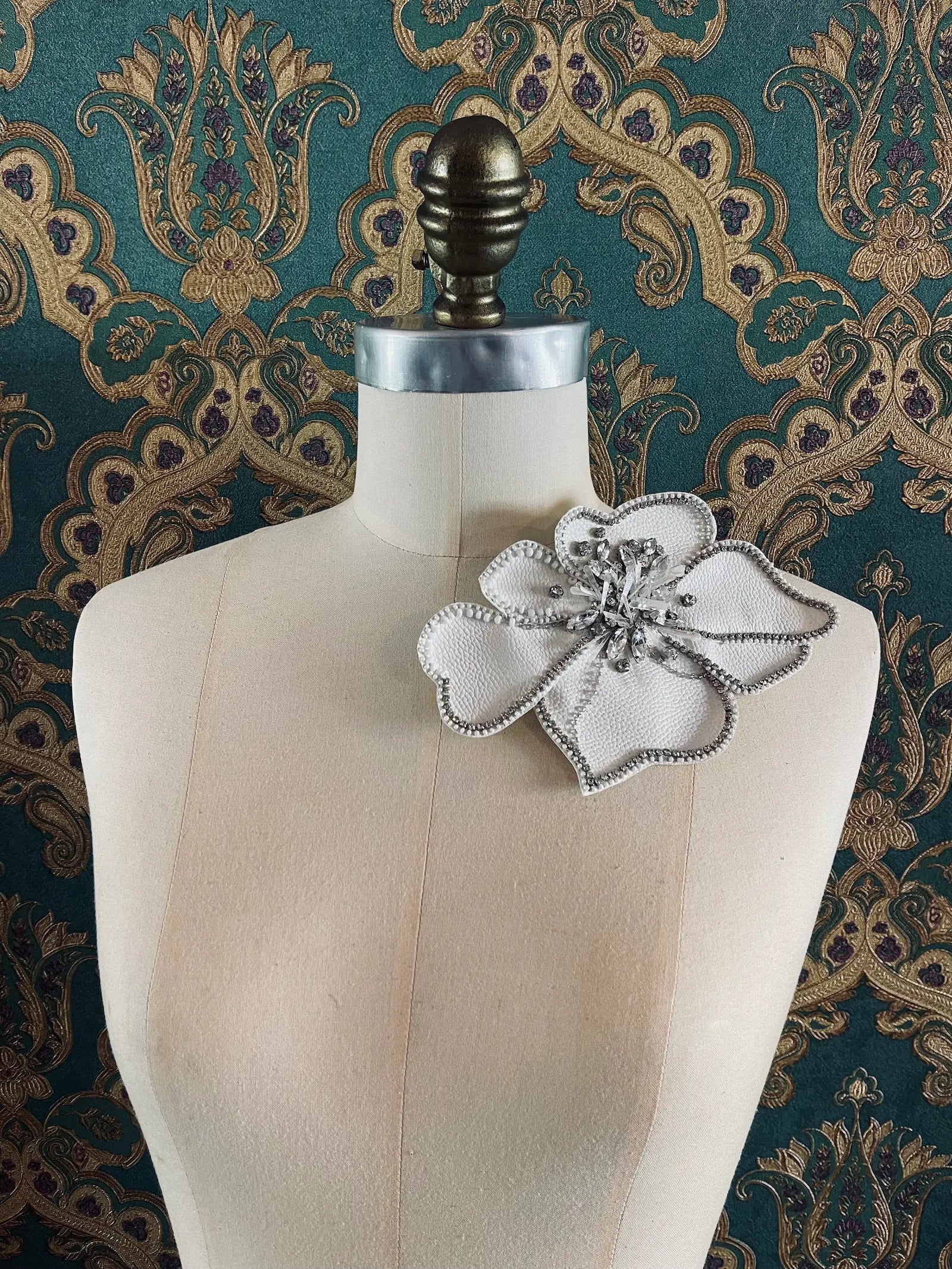Broochella Koi large white leather flower brooch with hand beaded crystal embellishment. Pictured on a mannequin. The background is green and gold damask wallpaper