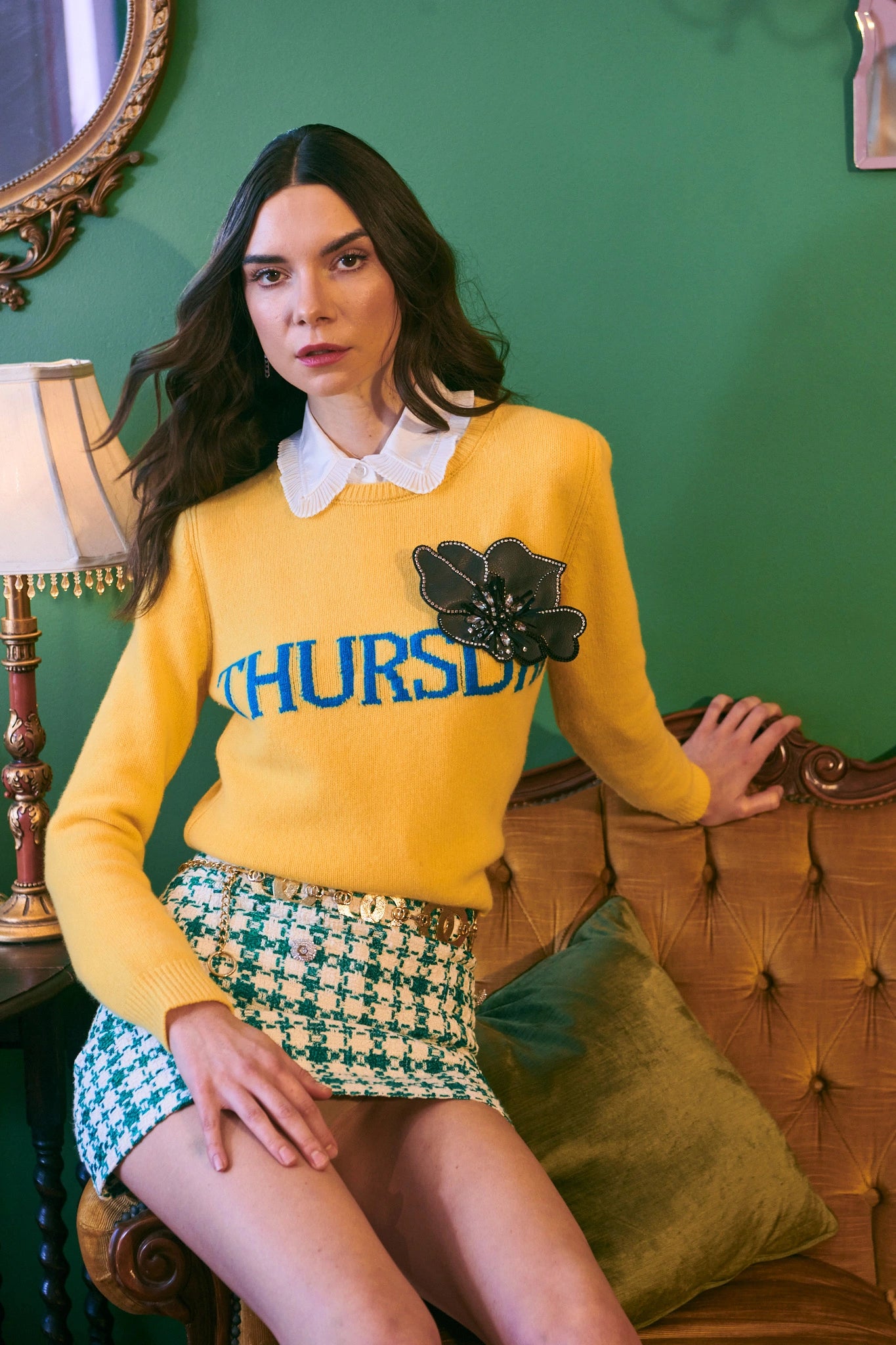 Dark haired model wearing the Broochella Koi large black flower brooch. She i leaning against a chair and is wearing a yellow jumper and a green mini skirt. The background is green.