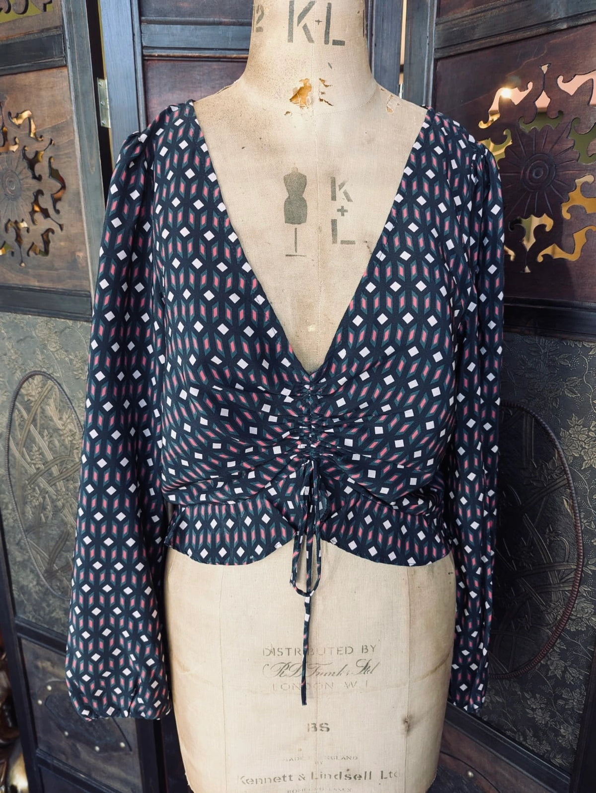 Broochella Kookai Long sleeve, silk printed blouse – Size 38. It is black, with a pink and white geometric print. There is a drawstring at the centre front of the top. It is pictured on a mannequin. The background is an ornate, dark wood room divider