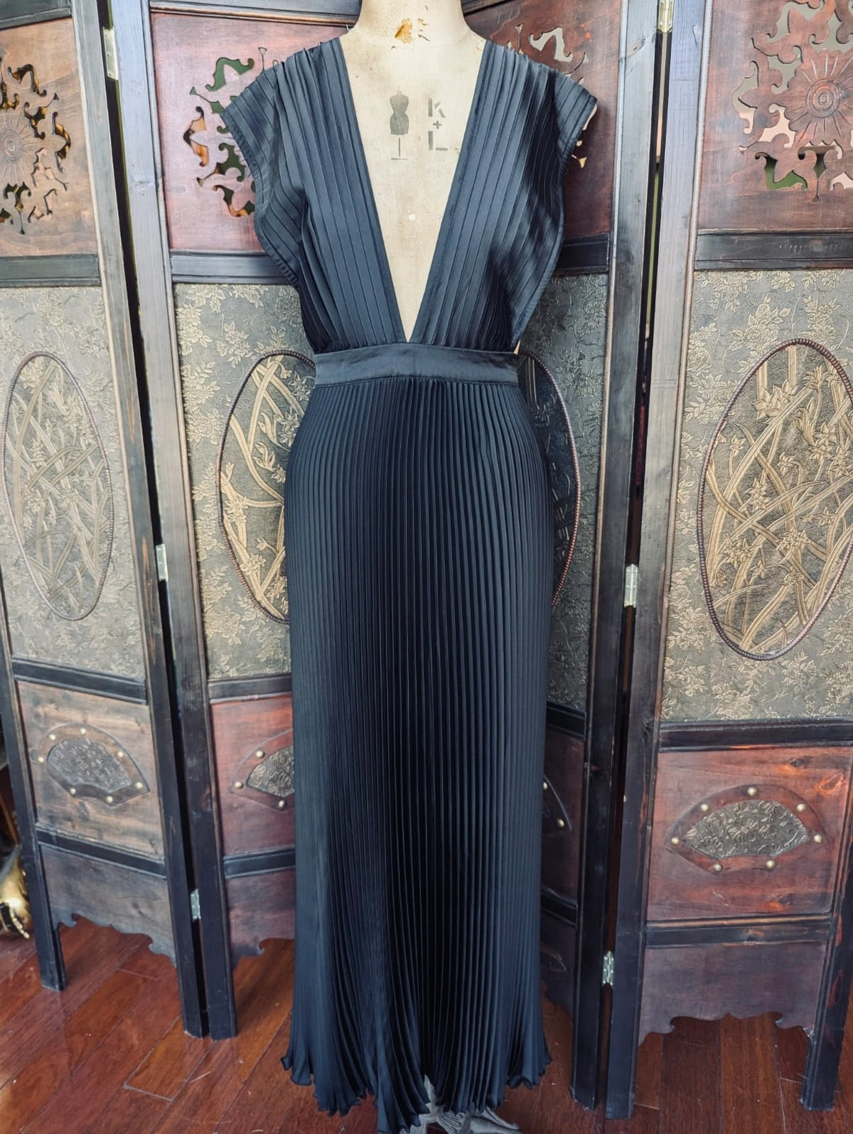 A Broochella L'idee black pleated maxi dress with a deep V-neckline and cap sleeves, displayed on a mannequin in front of a decorative folding screen.