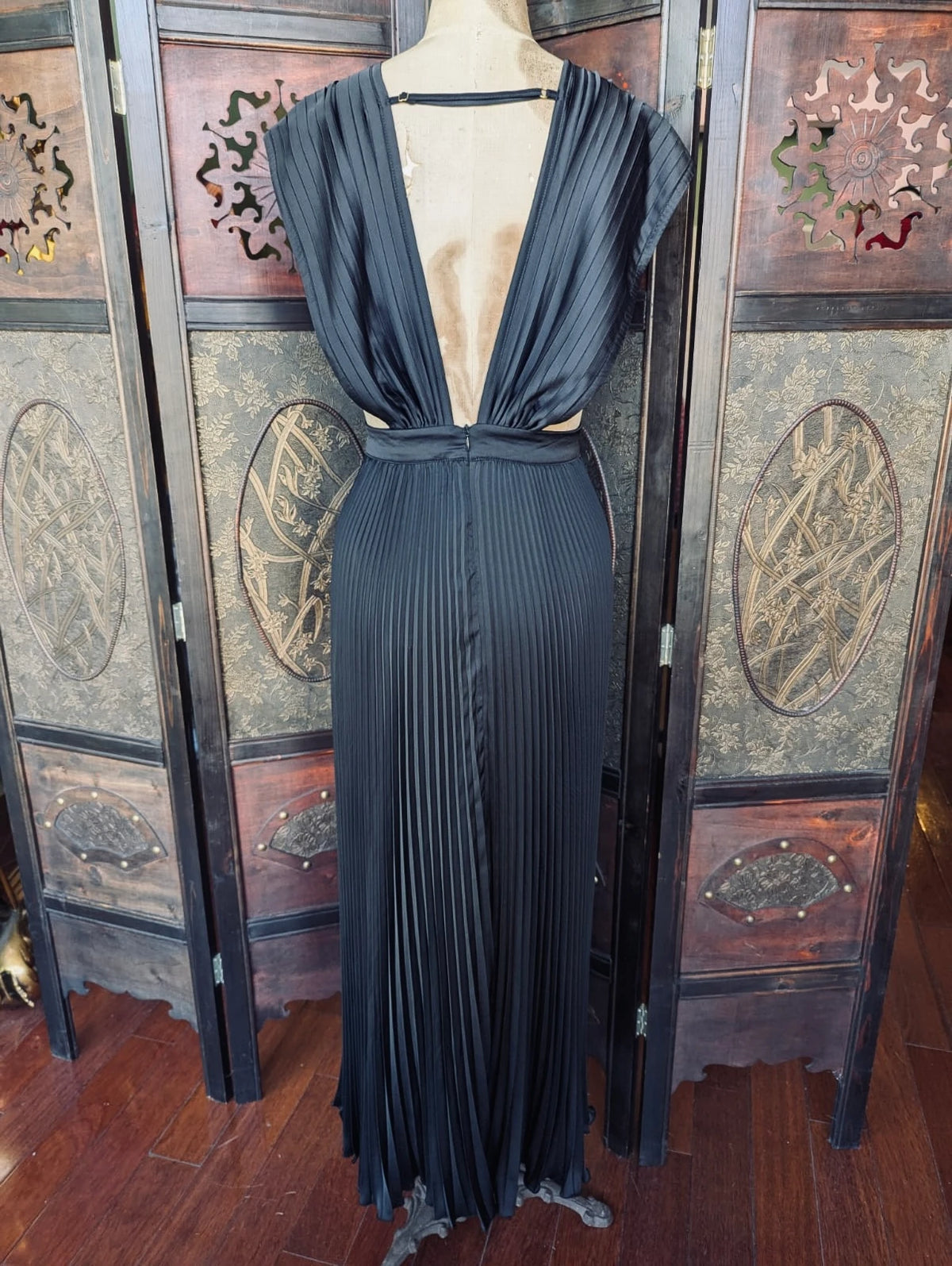 A Broochella L'idee black pleated maxi dress with a deep V-neckline back and cap sleeves, displayed on a mannequin in front of a decorative folding screen.