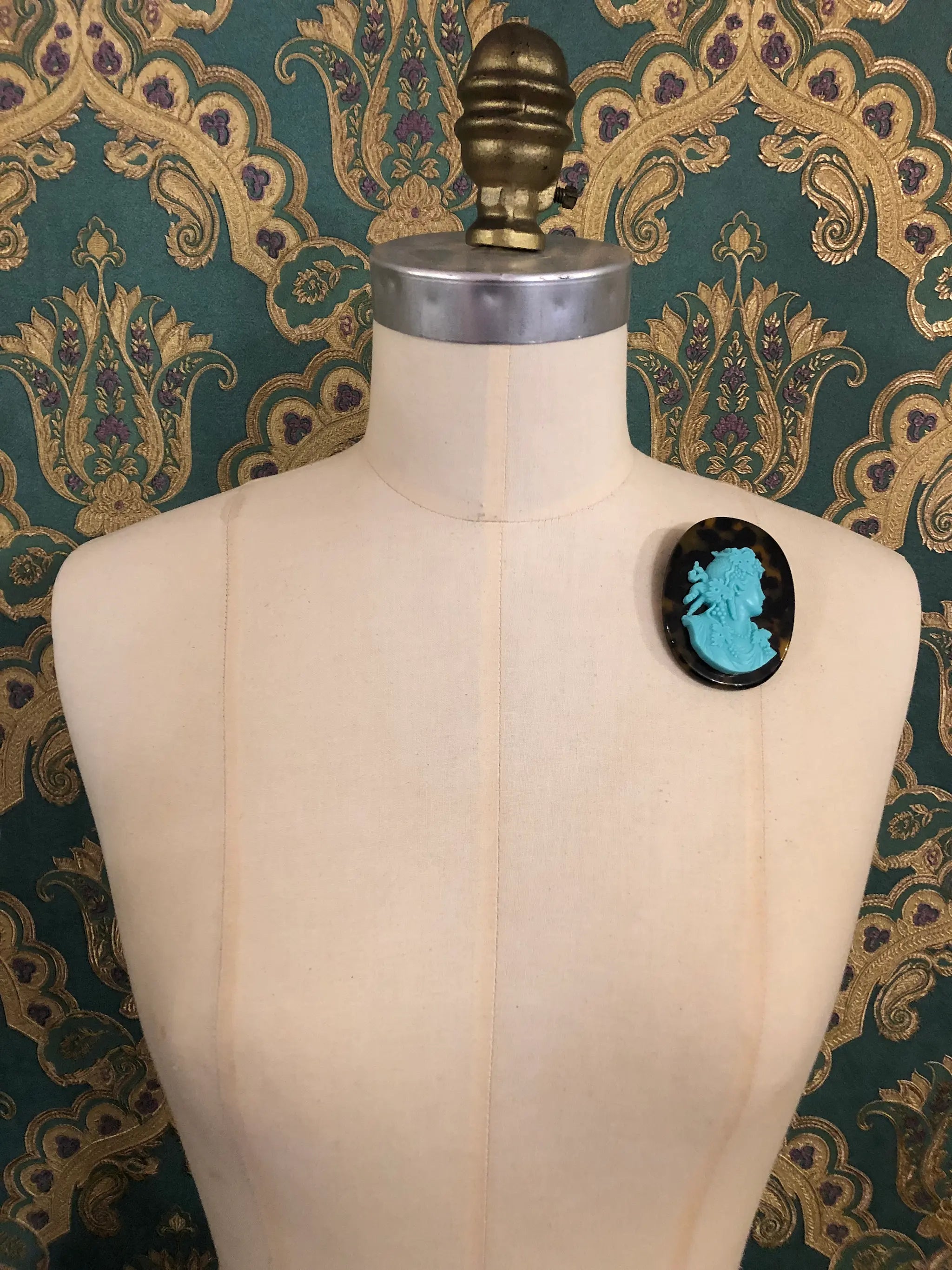Broochella La Bella Figura Cameo brooch. An aqua cameo on a tortoiseshell base,  displayed on a cream dress form against a dark teal and gold patterned fabric backdrop.