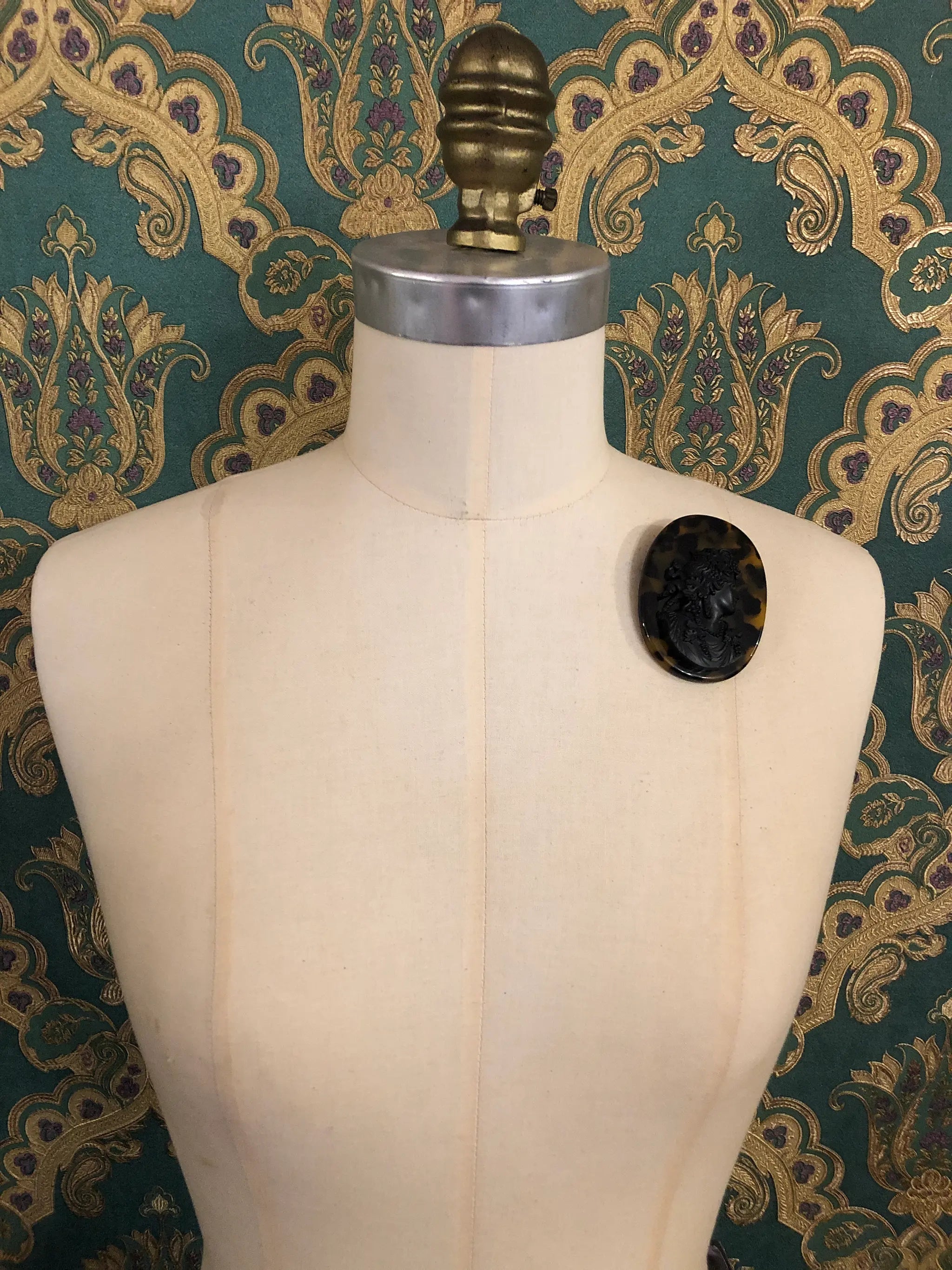 Broochella La Bella Figura Cameo brooch. A black cameo on a tortoiseshell base,  displayed on a cream dress form against a dark teal and gold patterned fabric backdrop.