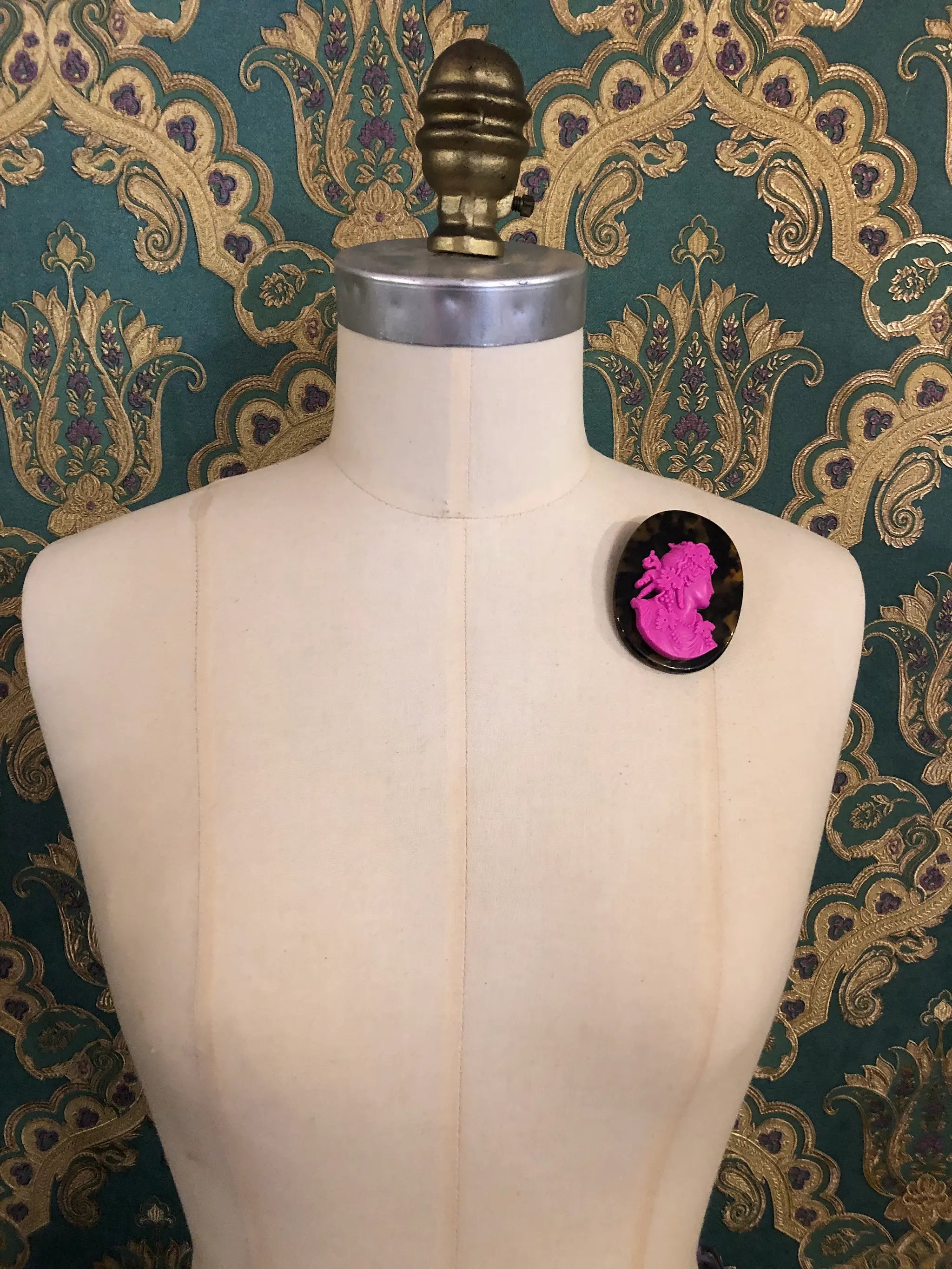 A Broochella La Bella Figura cameo Tortoiseshell brooch with a bright pink classical profile bust, displayed on a cream-coloured dress form against a dark teal and gold patterned wallpaper background.