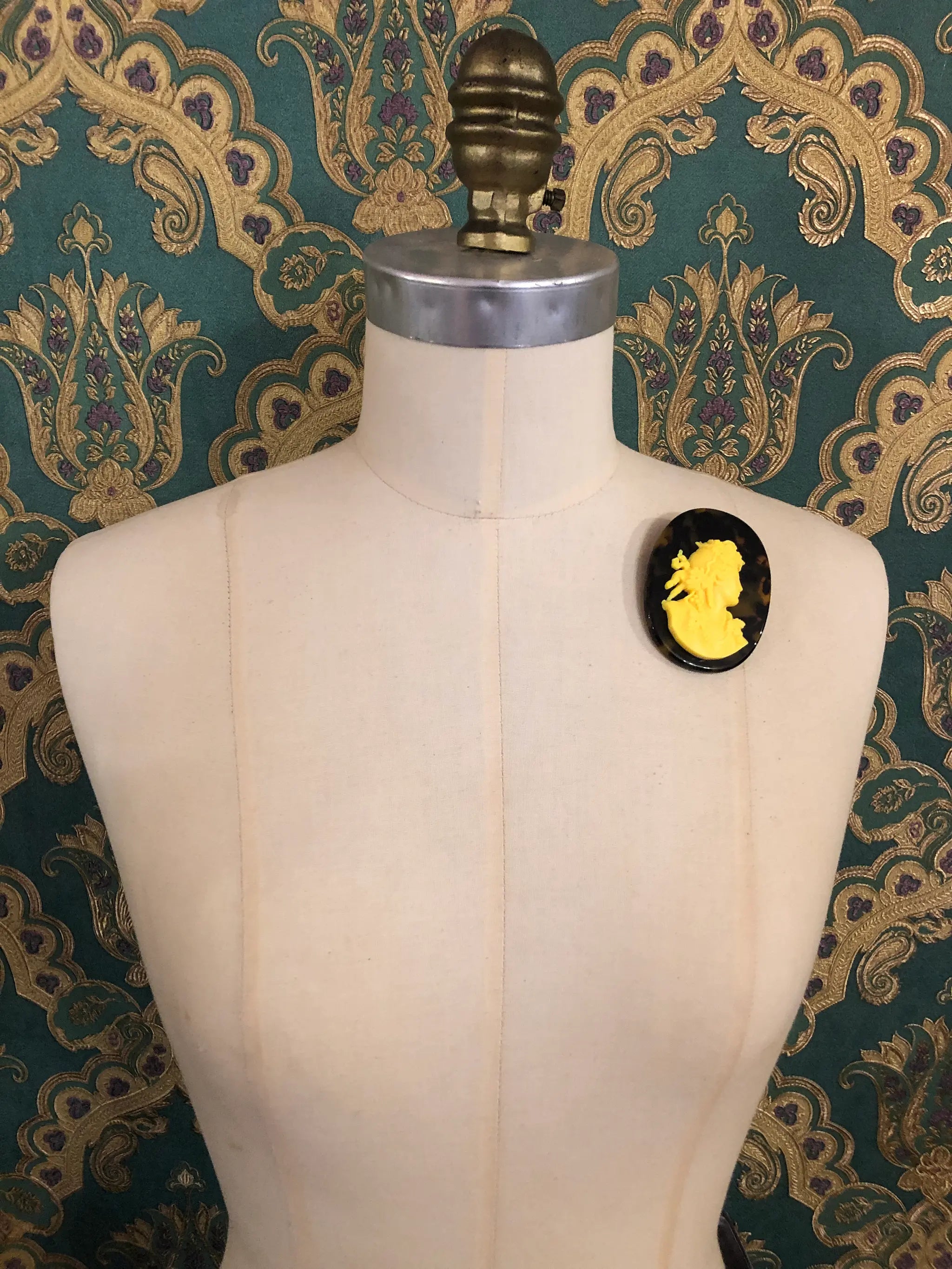 A Broochella la bella figura Tortoiseshell cameo oval brooch with a yellow classical profile silhouette, displayed on a cream-coloured dress form against a teal and gold patterned wallpaper background.
