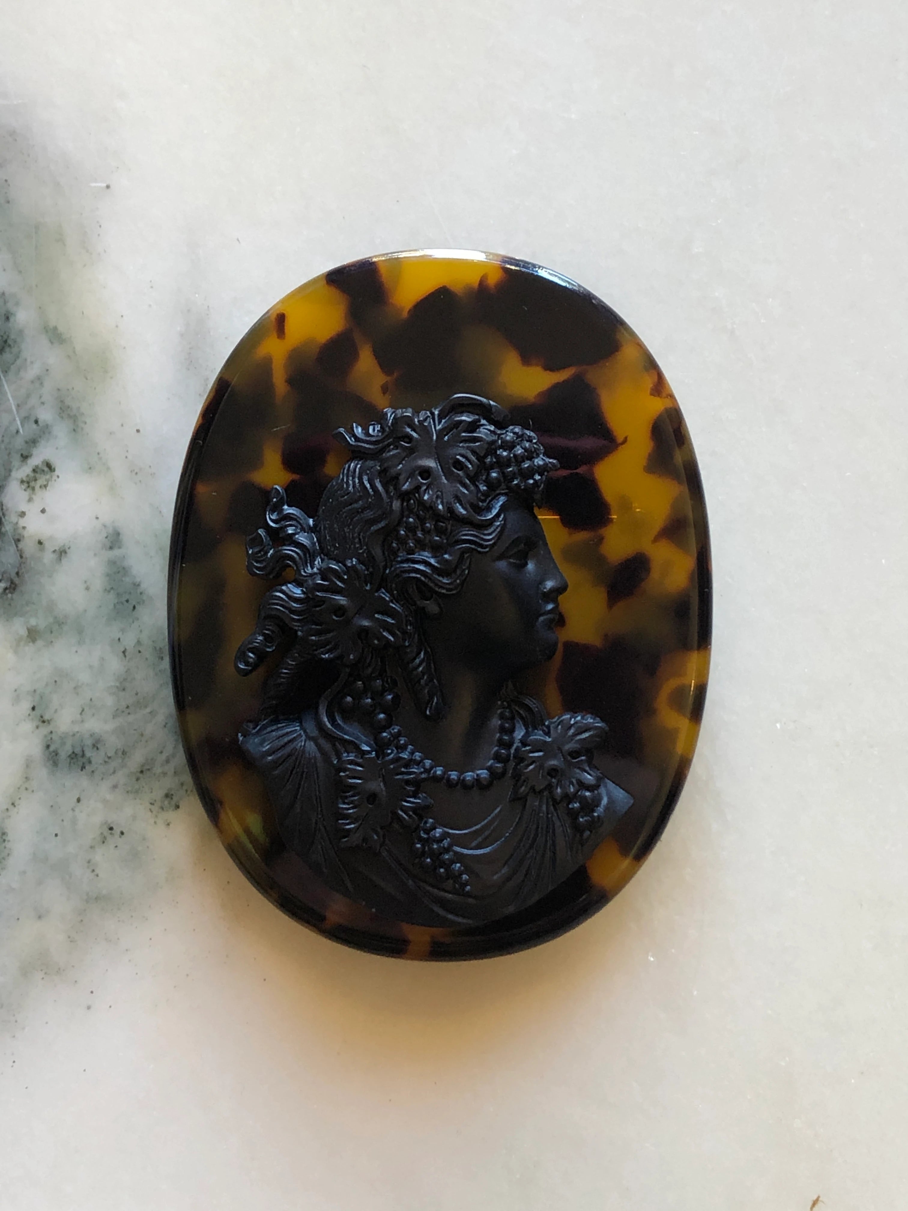 An oval Broochella La Bella Figura Cameo brooch featuring a classical profile portrait in dark carved stone against a mottled brown and amber tortoiseshell background, resting on a light marble surface.