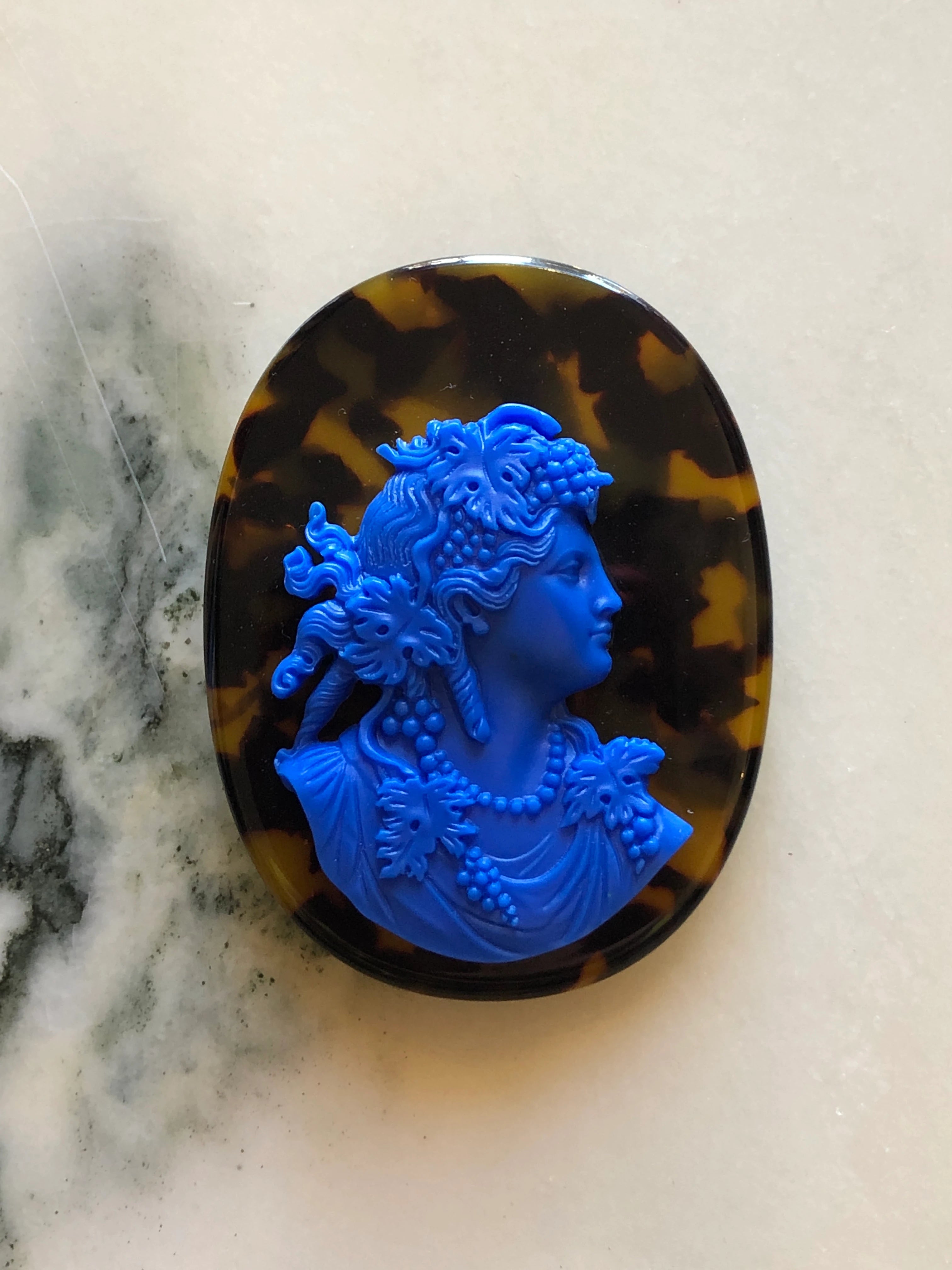 An oval Broochella La Bella Figura tortoiseshell cameo brooch with a bright blue carved profile portrait set into a dark tortoiseshell-patterned border, photographed on a light marble-like surface