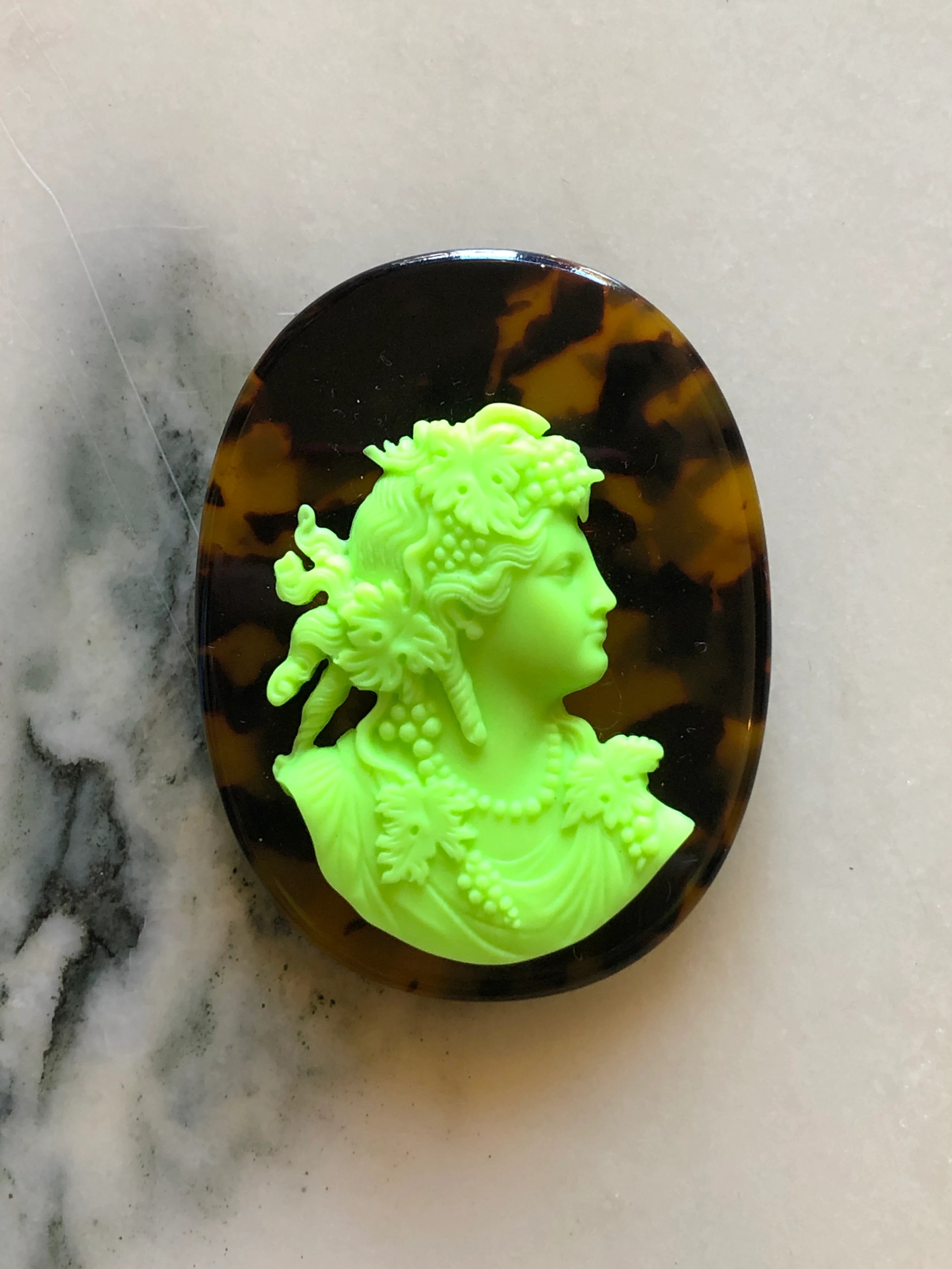 A Broochella La Bella Figura oval cameo brooch featuring a bright neon green classical profile bust of a woman with floral details, set against a dark brown and amber tortoiseshell-patterned background, displayed on a light marble surface.