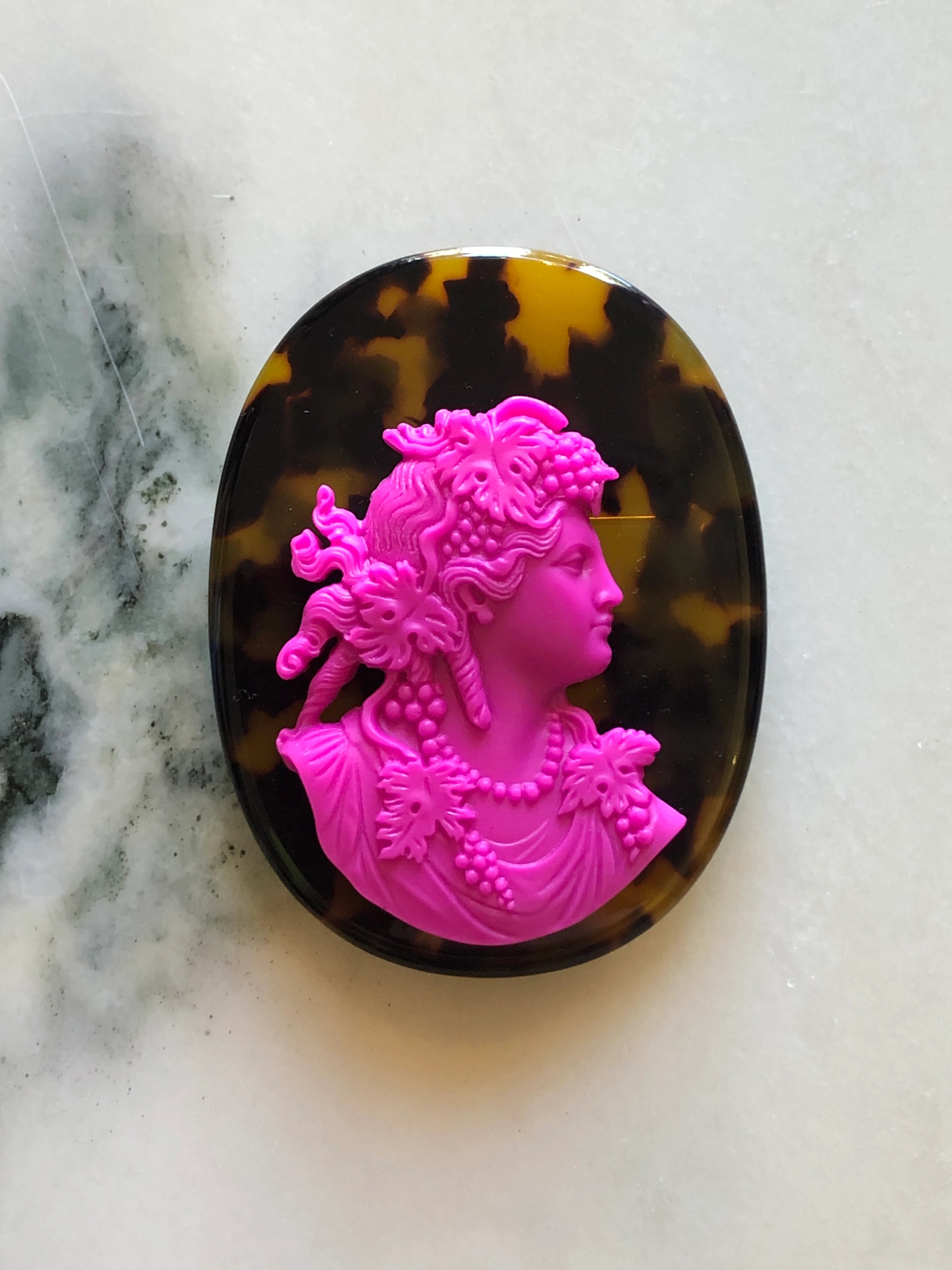 A Broochella La Bella Figura oval cameo brooch featuring a bright pink classical profile bust of a woman with floral details, set against a dark brown and amber tortoiseshell-patterned background, displayed on a light marble surface.