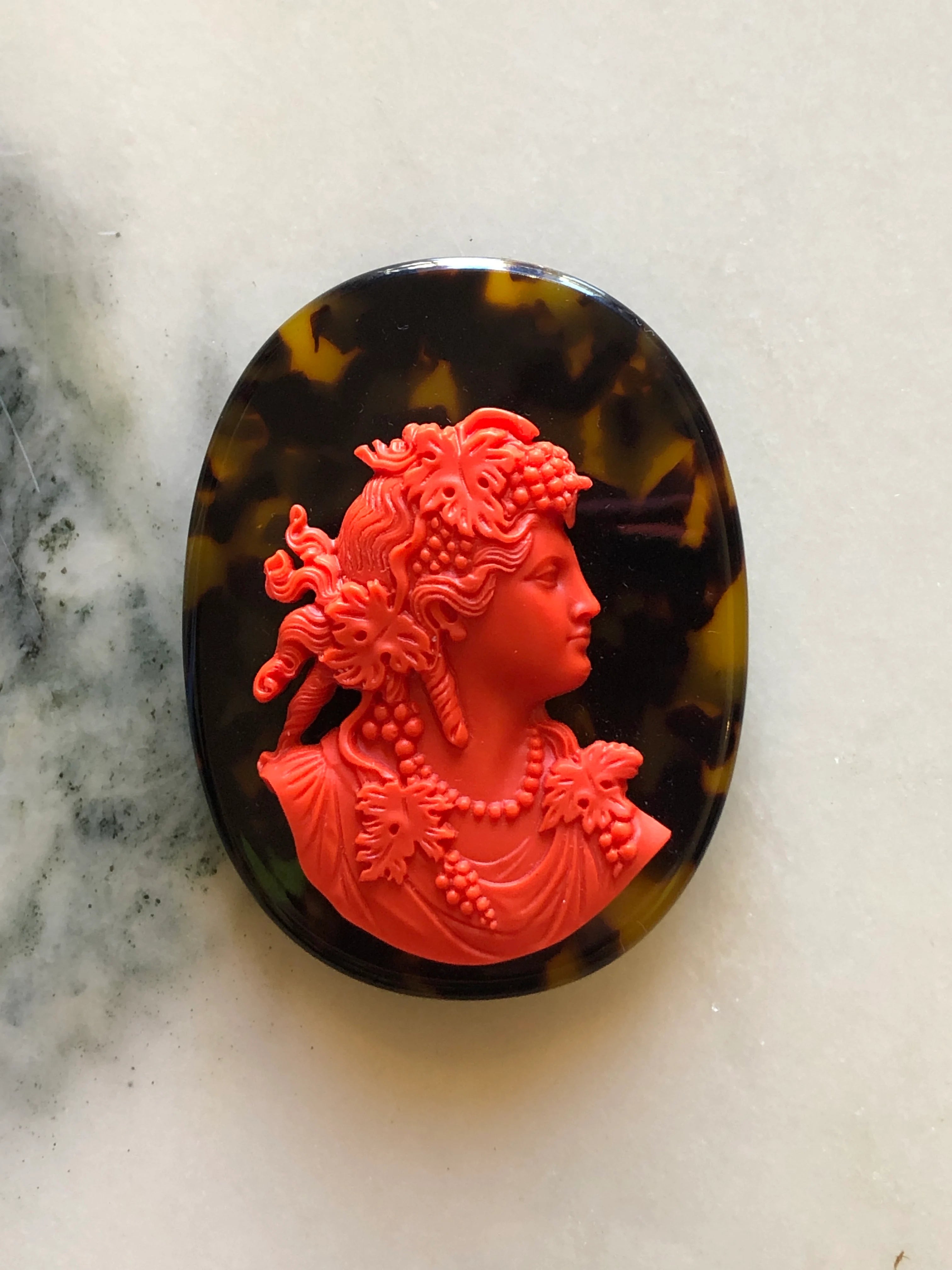 An oval Broochella La Bella Figura Tortoiseshell cameo brooch featuring a bright red classical bust relief set into a dark tortoiseshell background, photographed on a light marble-like surface.