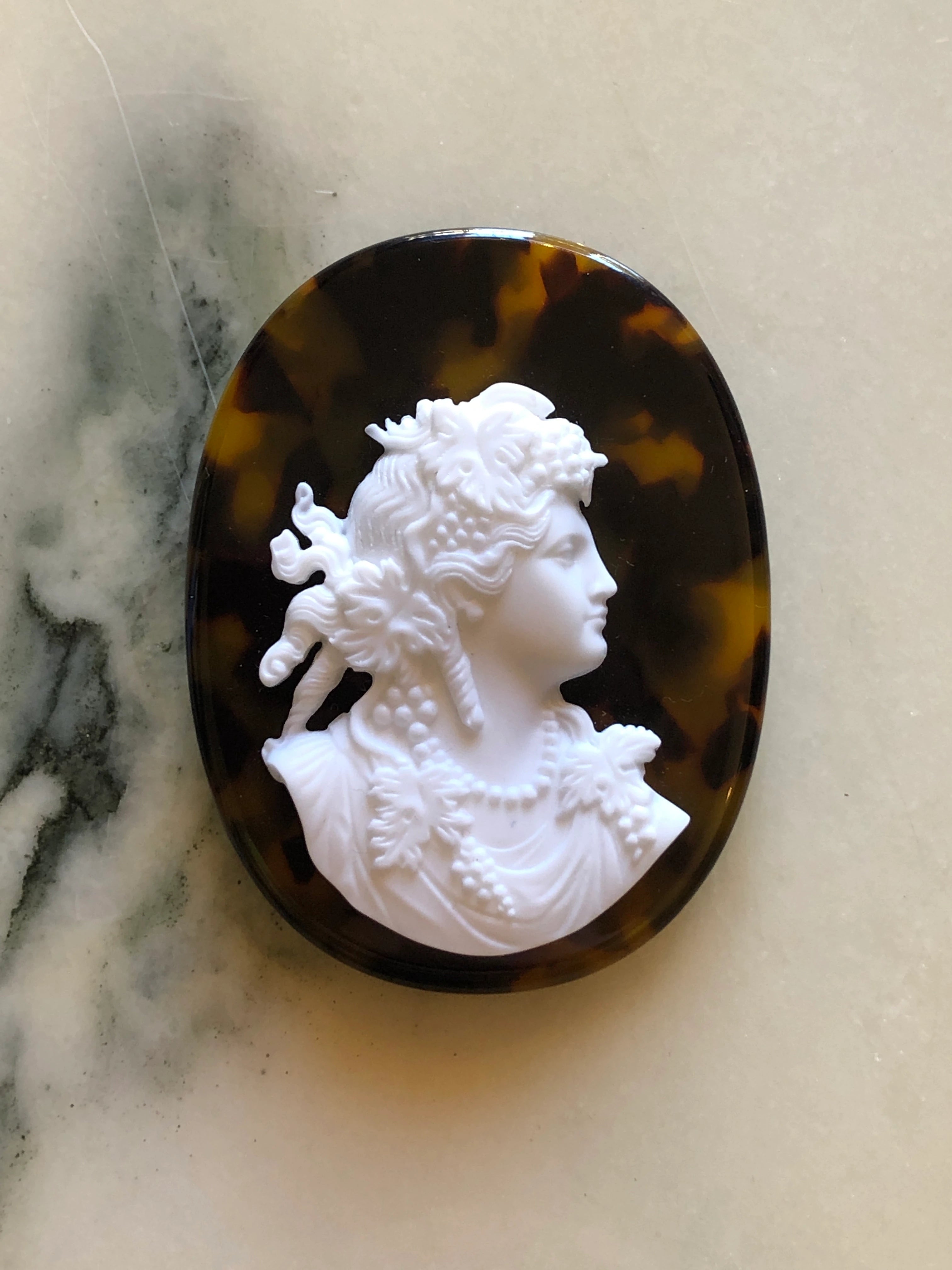 A Broochella La Bella Figura oval cameo brooch featuring a white classical profile bust of a woman with floral details, set against a dark brown and amber tortoiseshell-patterned background, displayed on a light marble surface.