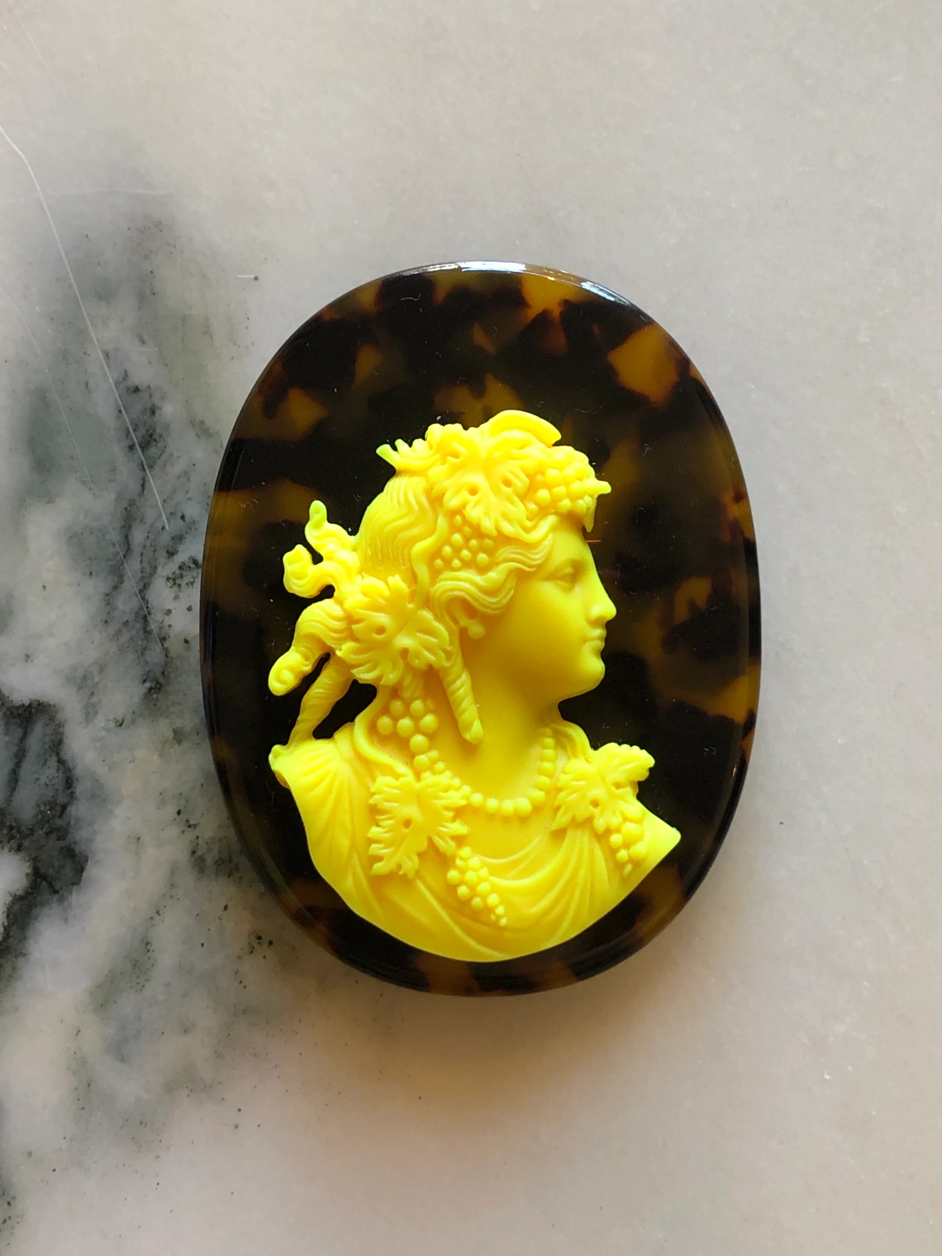 An oval Broochella Men's La Bella Figura Tortoiseshell Cameo Brooch featuring a bright yellow classical bust relief of a woman in profile, set against a dark brown and black tortoiseshell background, displayed on a light marble surface.