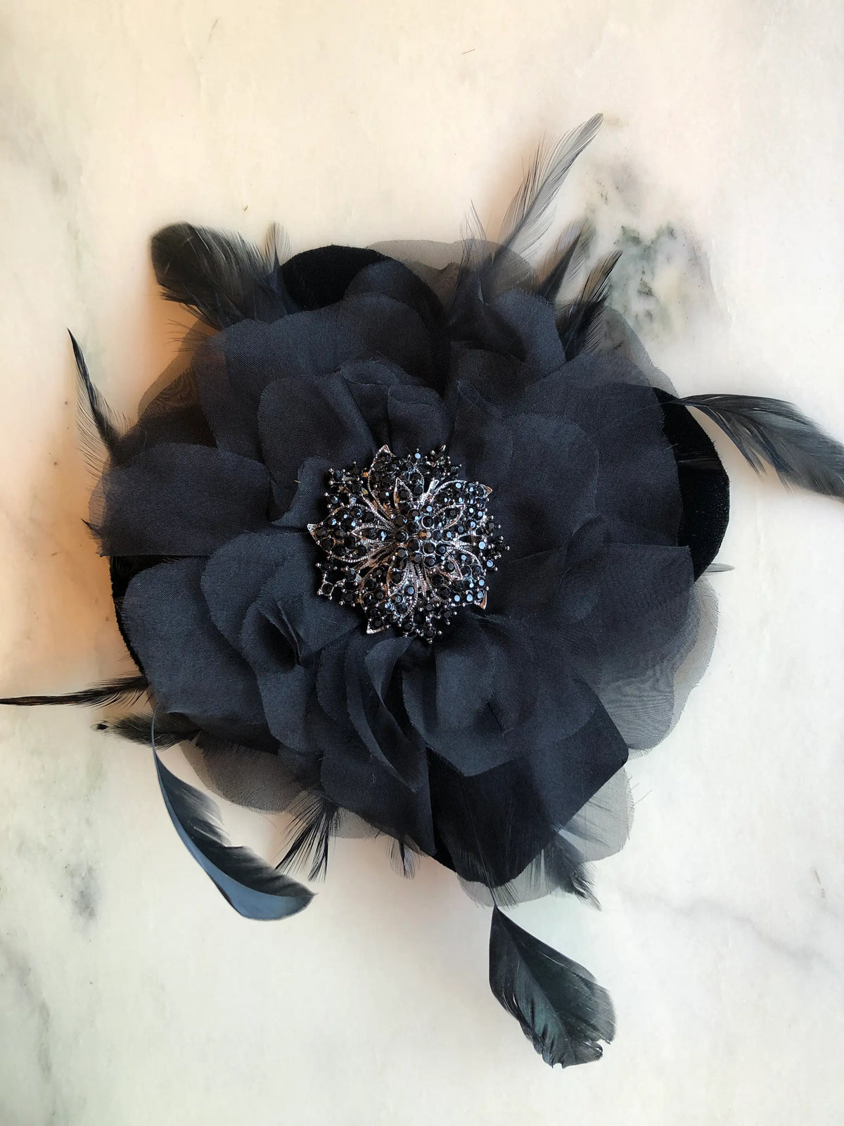 Broochella Men's La Contessa Flower Brooch Pin in black, featuring layered fabric petals, delicate black feathers and an ornate black crystal centre, photographed on a white marble background with soft grey veining.