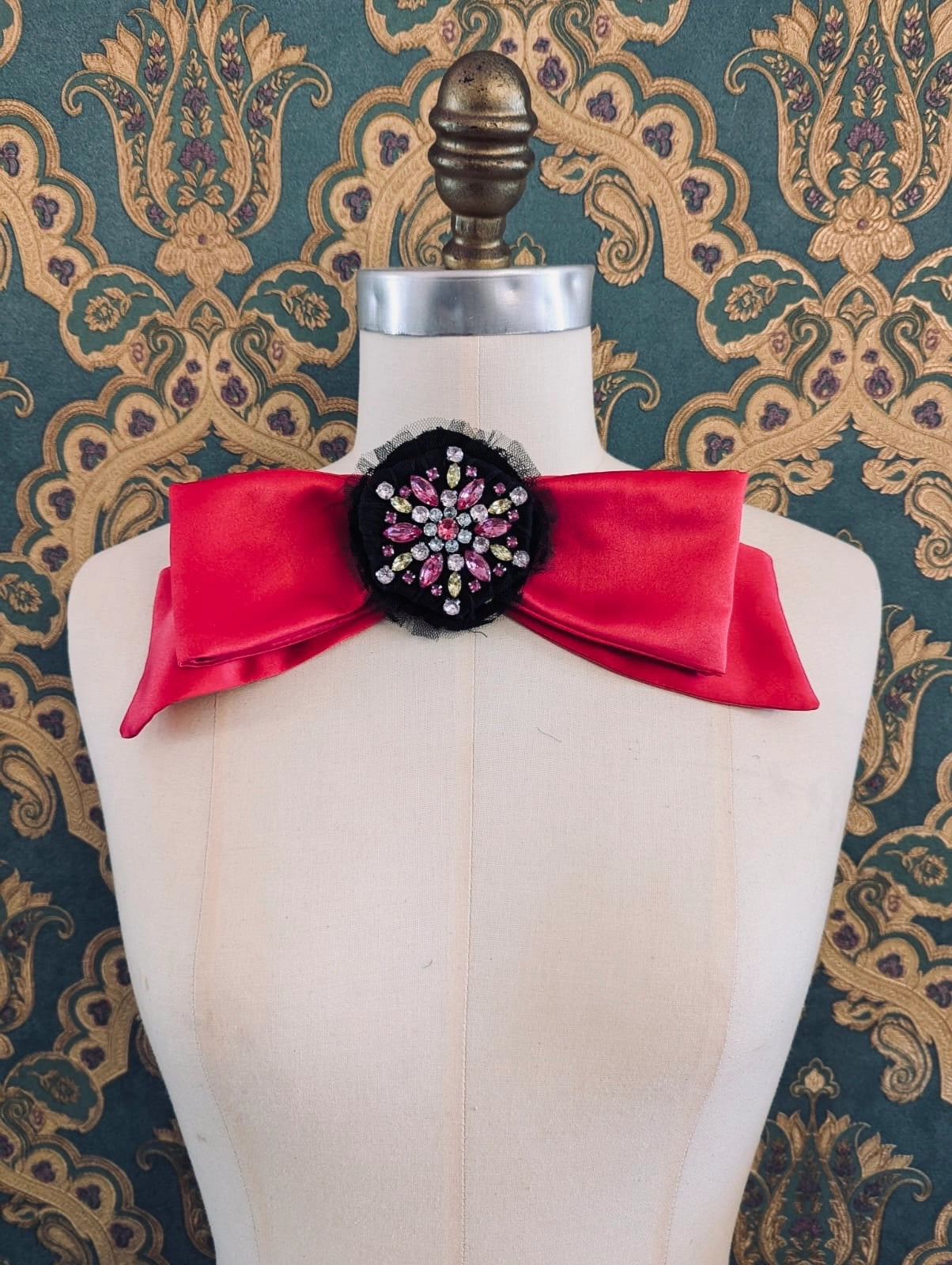 Broochella la dolce vita handmade pink satin bow. It had a black and pink brooch embellishment at the centre if the bow.  It is pinned to a mannequin. The background is green and gold damask wallpaper.
