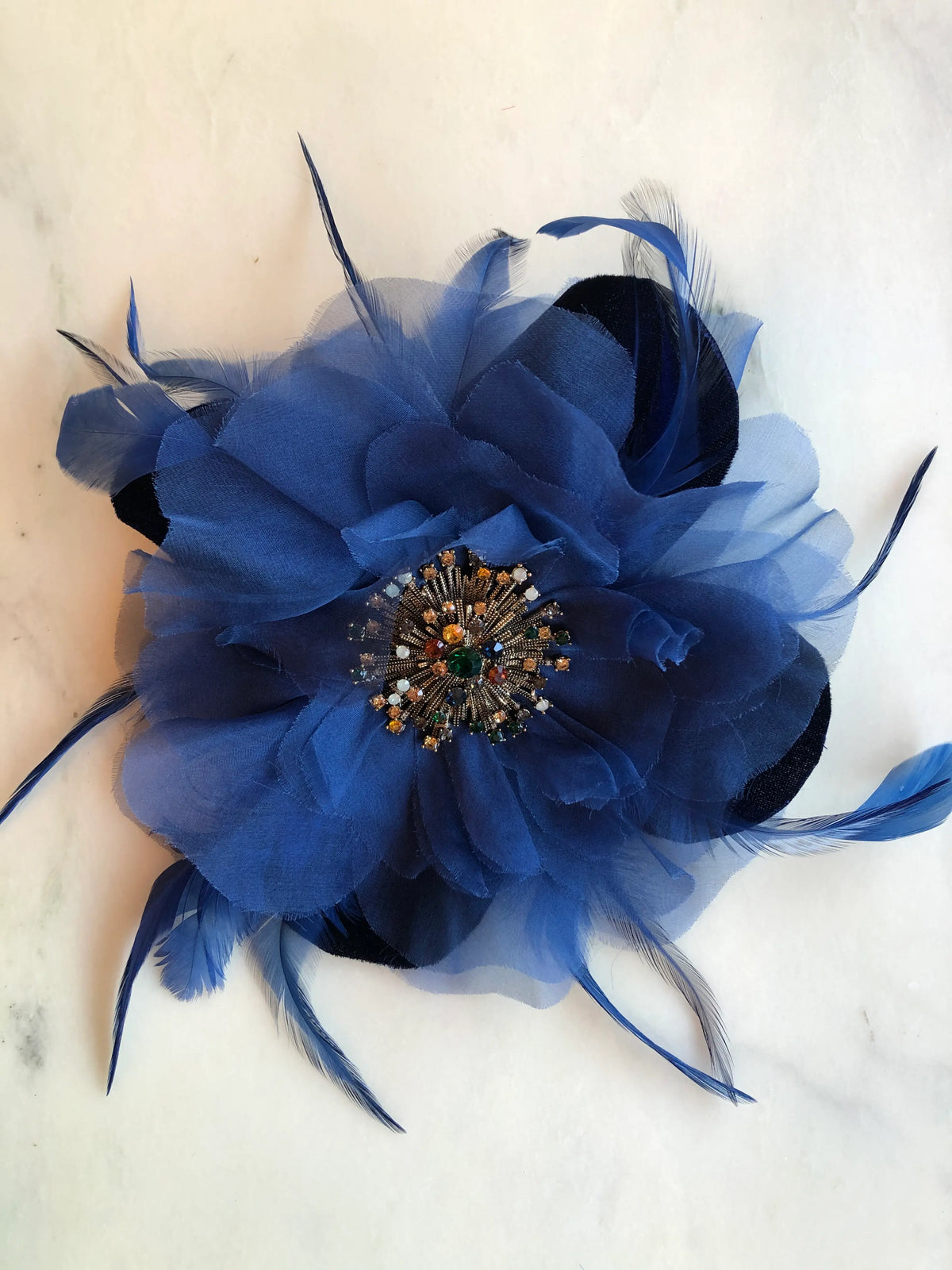 Broochella La Duchessa Flower Brooch Pin in deep royal blue, featuring layered sheer fabric petals, sweeping blue feathers and an ornate multicoloured jewelled centre, photographed on a white marble background with soft grey veining.