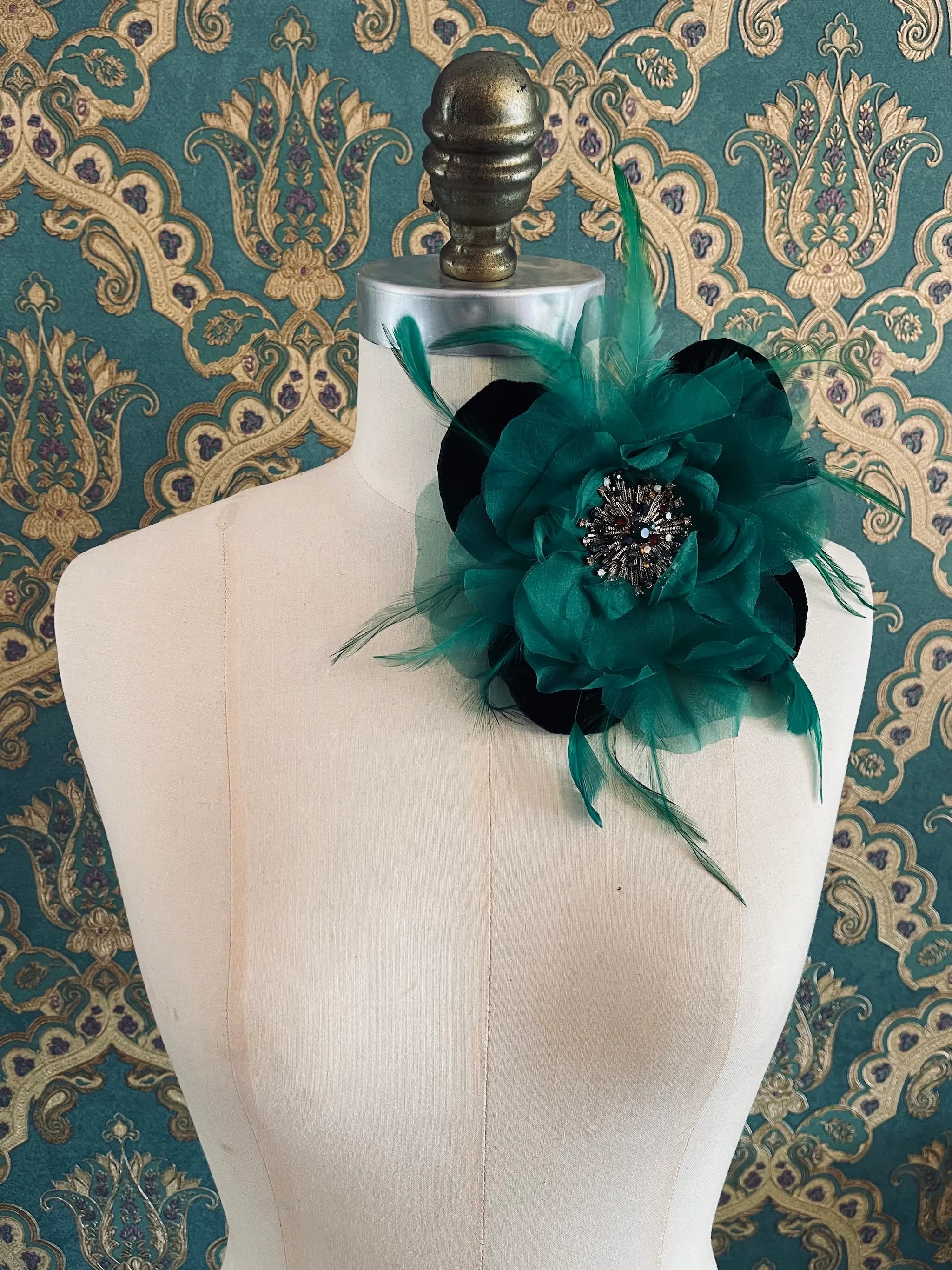 Broochella la duchessa green handmade fabric and feather flower brooch with centre embellishment. Pictured on a mannequin. The background is green and gold damask wallpaper