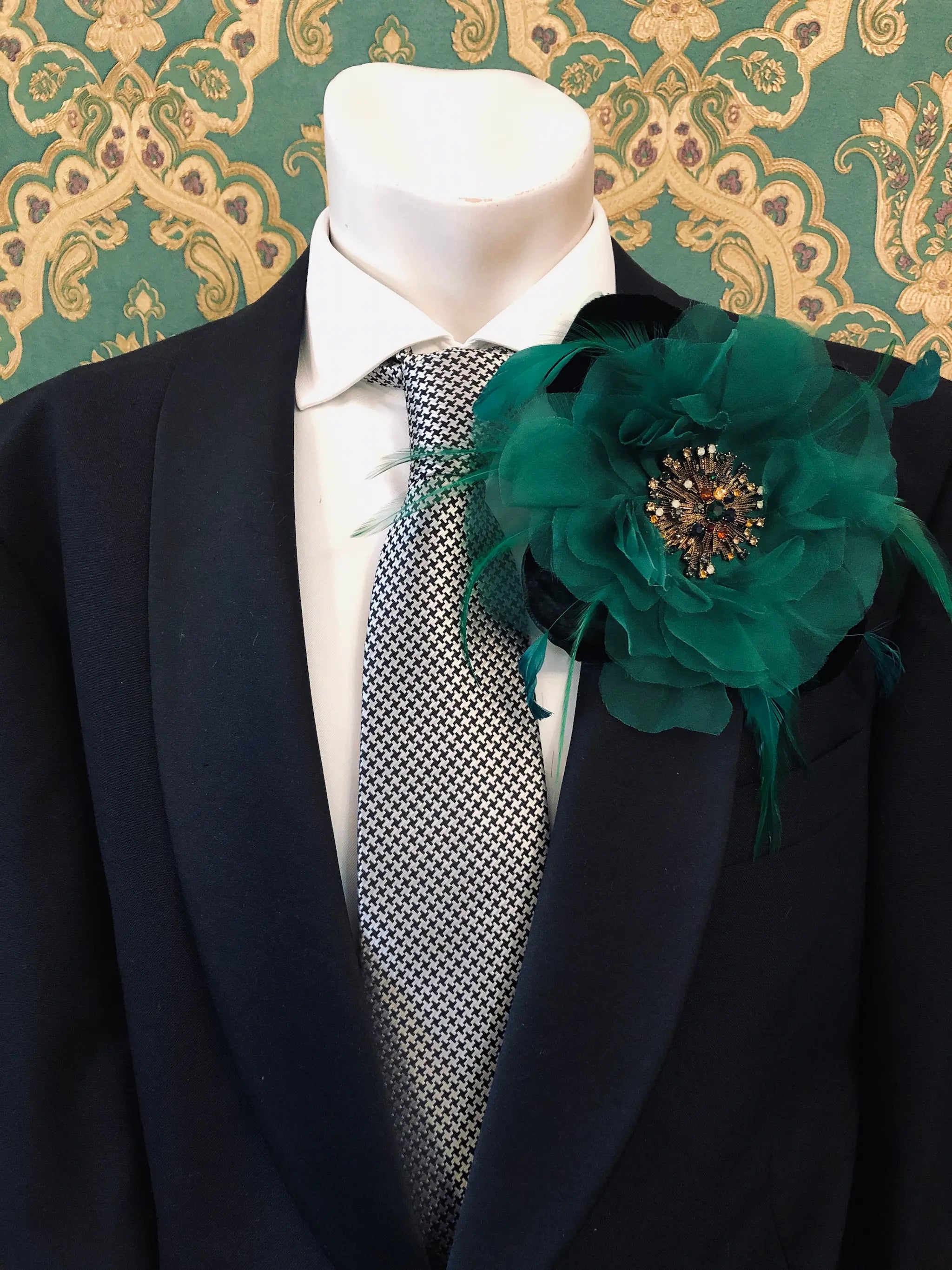A black suit jacket on a mannequin torso, styled with a white dress shirt, a grey and white patterned tie, and a Broochella Men's la Duchessa large emerald green fabric flower brooch with a jewelled centre, set against a green damask wallpaper background.