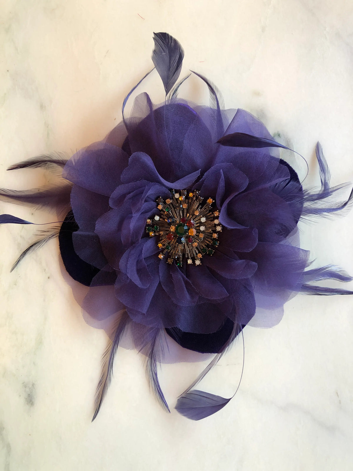 Broochella Men's La Duchessa Flower Brooch Pin in deep purple, featuring layered sheer fabric petals, delicate sweeping feathers and an ornate multicoloured jewelled centre, photographed on a white marble background with soft grey veining.