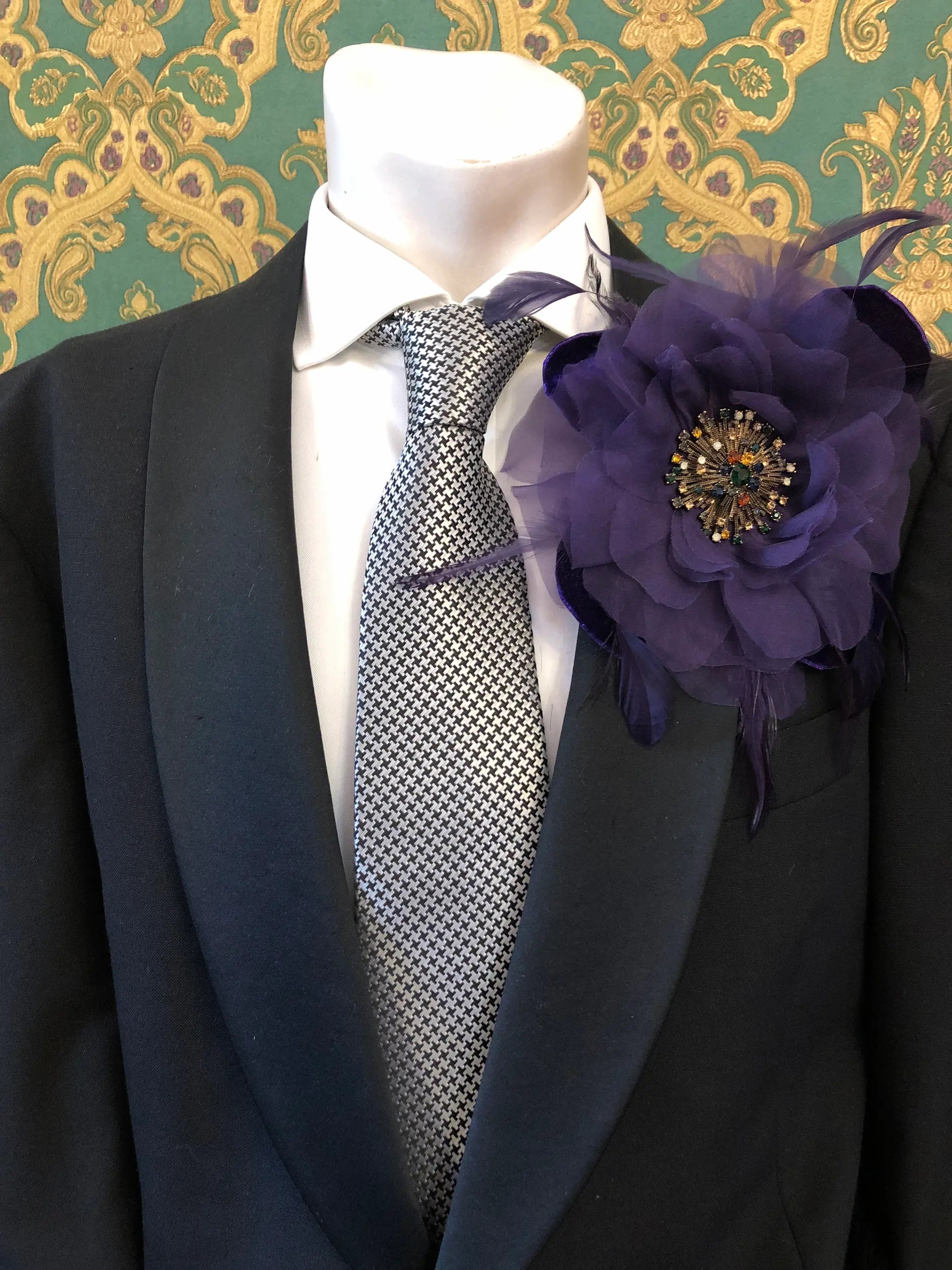 A black suit jacket on a mannequin torso, styled with a white dress shirt, a grey and white patterned tie, and a Broochella Men's la Duchessa large purple fabric flower brooch with a jewelled centre, set against a green damask wallpaper background.