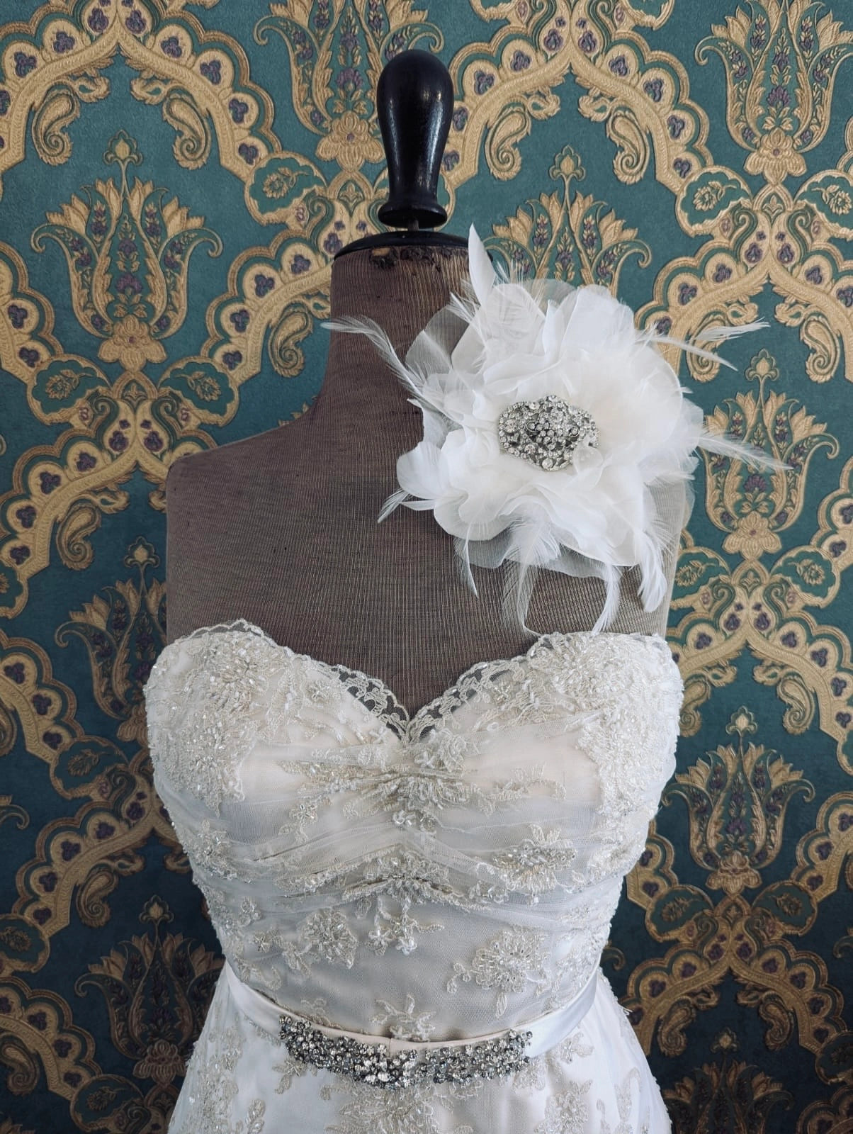 Broochella la sposa white velvet, organza and feather brooch with a silver crystal brooch embellishment at the centre. Pictured on a mannequin that is wearing a white, beaded strapless wedding dress. The background is green and gold damask wallpaper. 