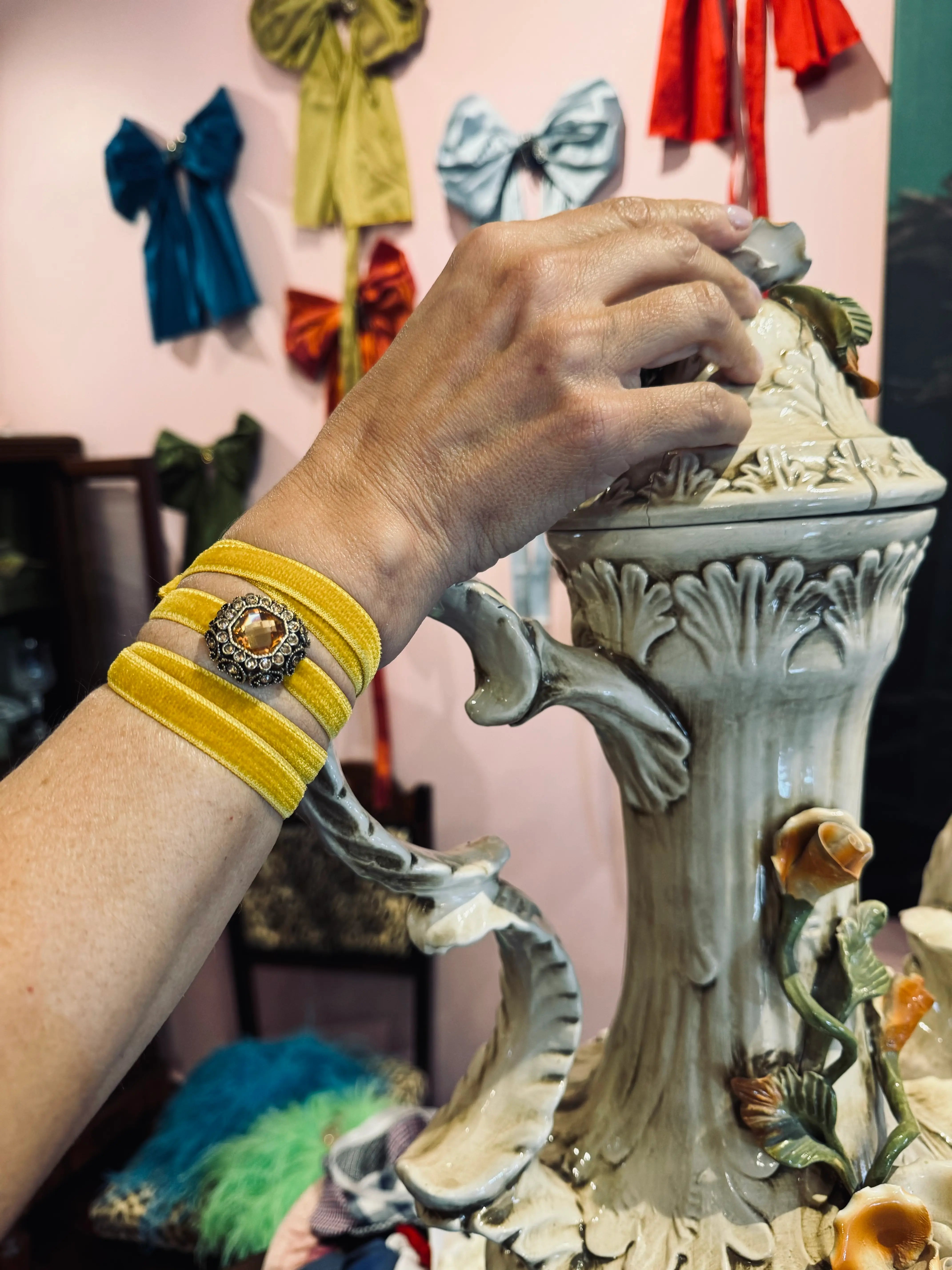 A close-up of a person's hand wearing a Broochella Lady Whistledown velvet choker with a central gold-toned brooch, reaching toward a decorative ceramic vase in a colorful room.