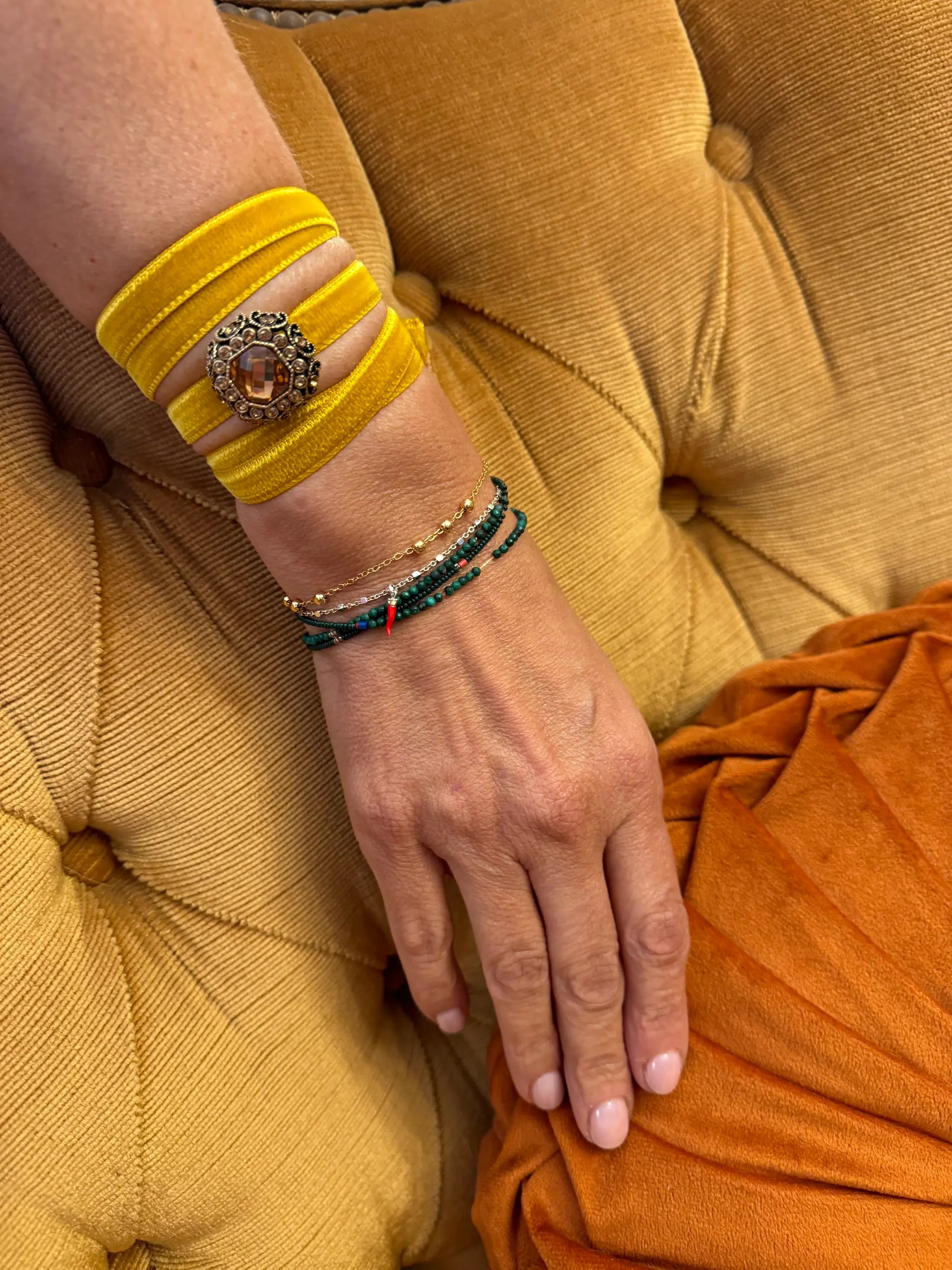 A close-up of a person's hand and wrist wearing the Broochella Lady Whistledown velvet choker, and teal-green beaded bracelets, resting on a mustard yellow tufted chair next to an orange fabric surface.