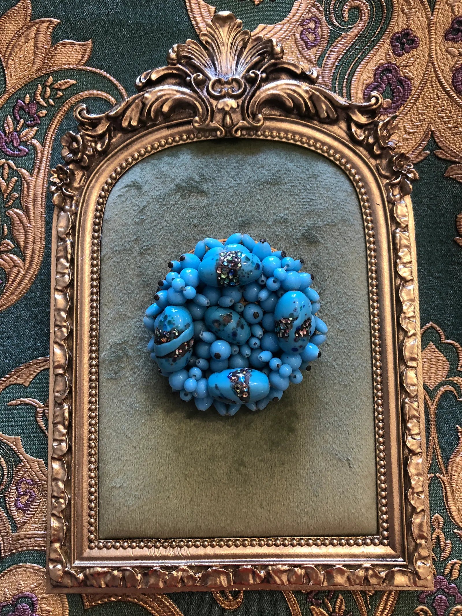A Broochella laguna Turchese brooch with circular cluster of bright turquoise blue beads with small silver-toned floral accents, displayed inside an ornate gold-framed mirror on a patterned green and gold fabric background.