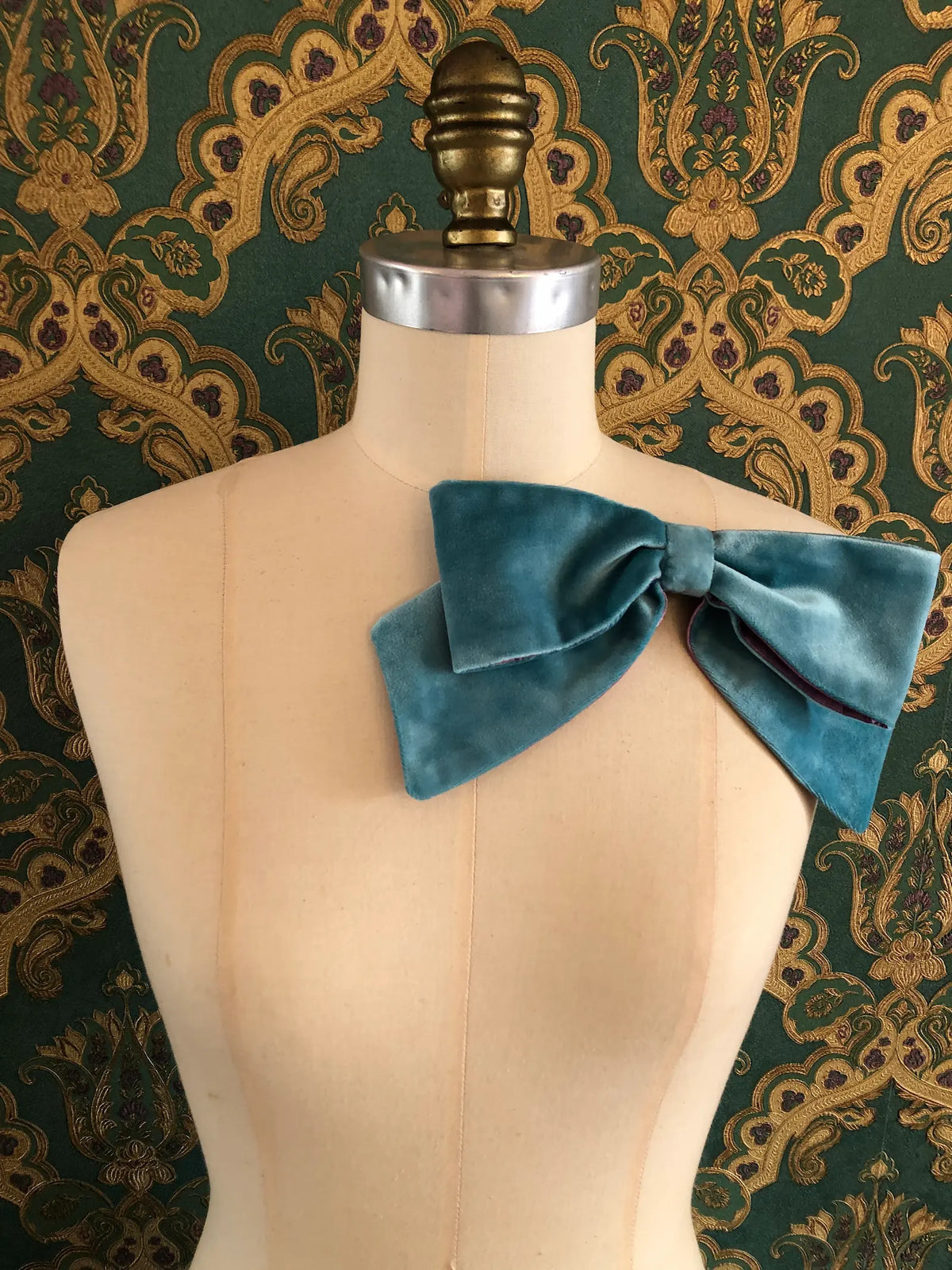 Broochella handmade Large Velvet Bow Brooch in aqua blue velvet with purple edging, styled on a cream mannequin against ornate green and gold wallpaper.