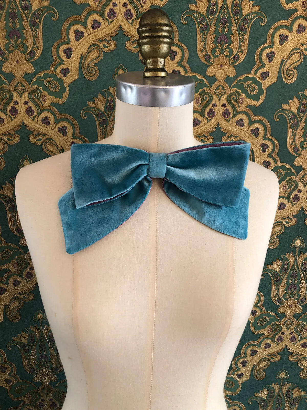 Broochella handmade Large Velvet Bow Brooch in aqua blue velvet with purple edging, styled on a cream mannequin against ornate green and gold wallpaper.