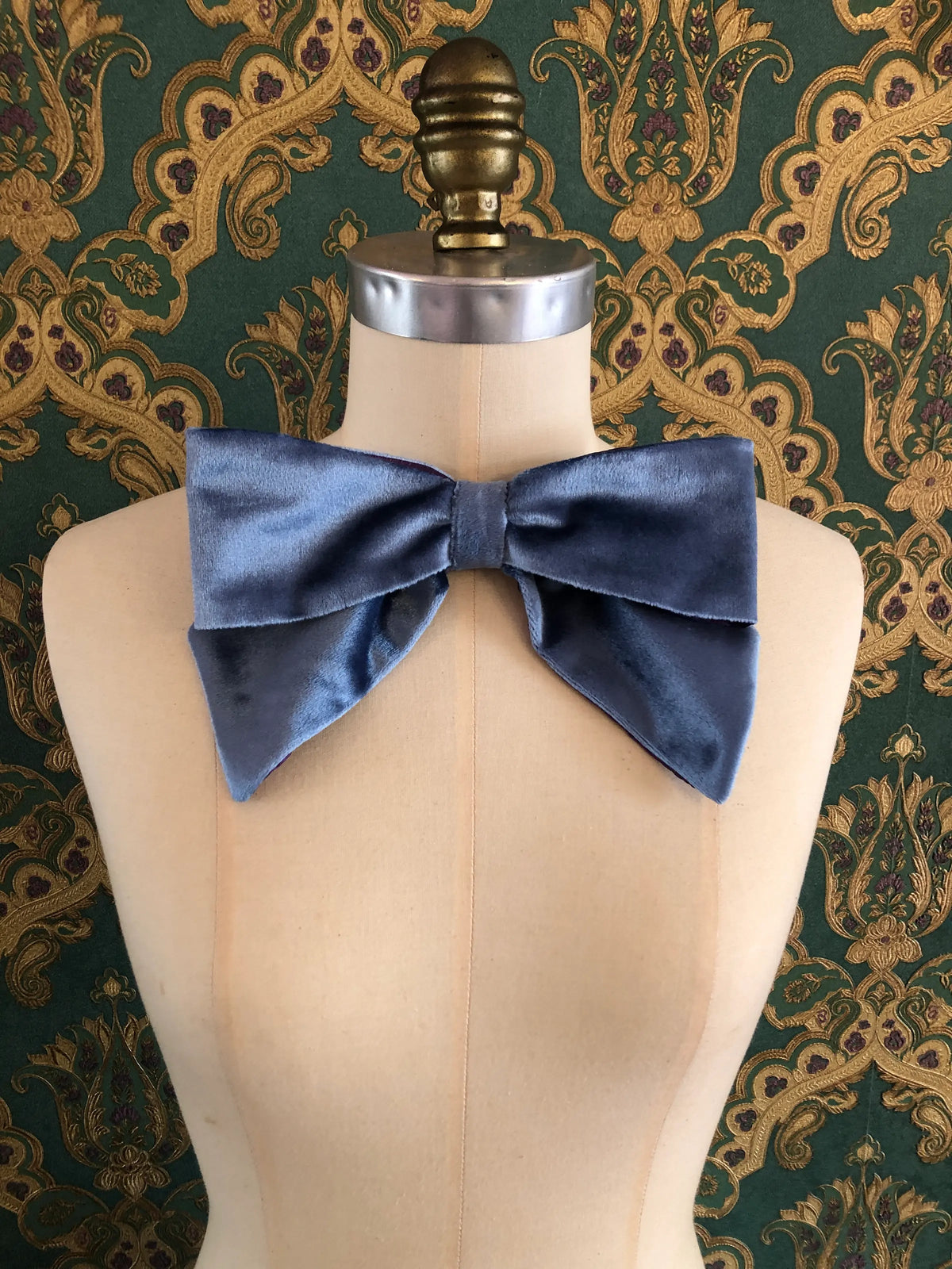 Broochella handmade Large Velvet Bow Brooch in soft blue velvet with contrasting purple edging, styled asymmetrically on a cream mannequin against ornate green and gold wallpaper.