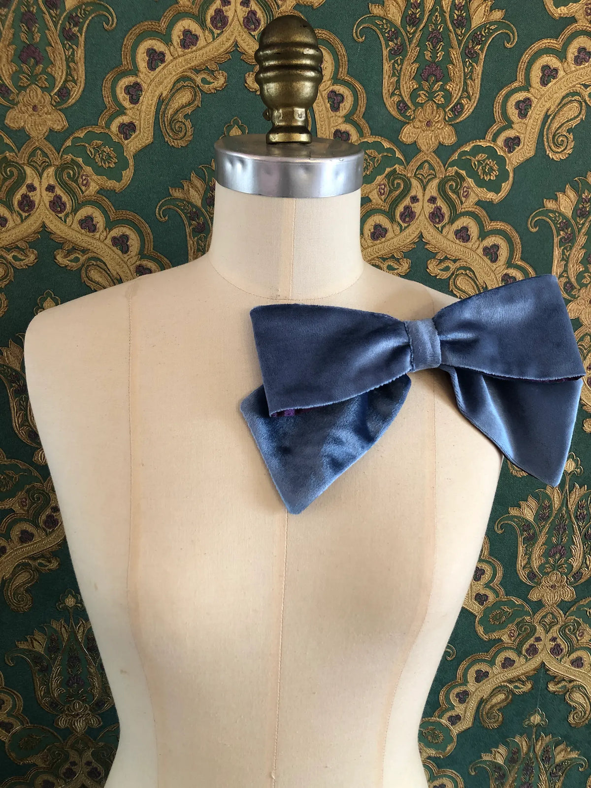Broochella handmade Large Velvet Bow Brooch in soft blue velvet with contrasting purple edging, styled asymmetrically on a cream mannequin against ornate green and gold wallpaper.