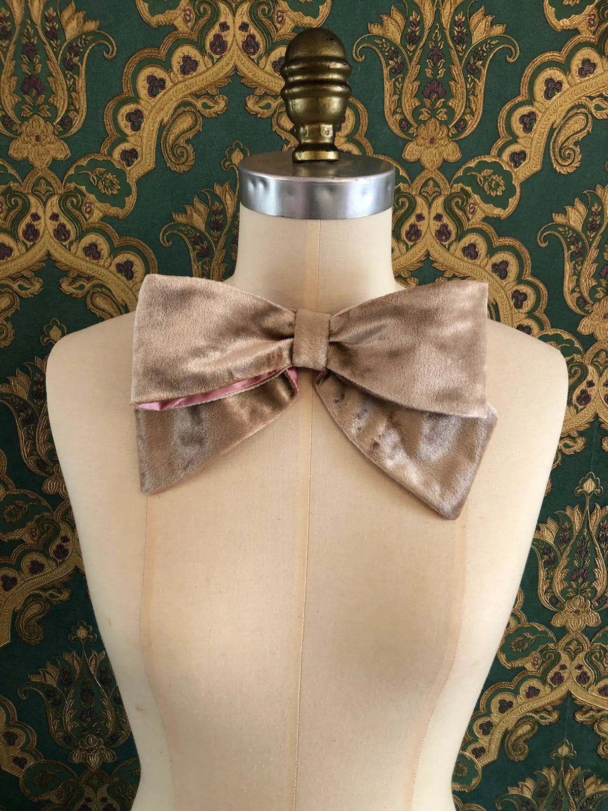 Broochella handmade Large Velvet Bow Brooch in luxurious champagne velvet with contrasting pink lining, styled asymmetrically on a cream mannequin against ornate green and gold wallpaper.