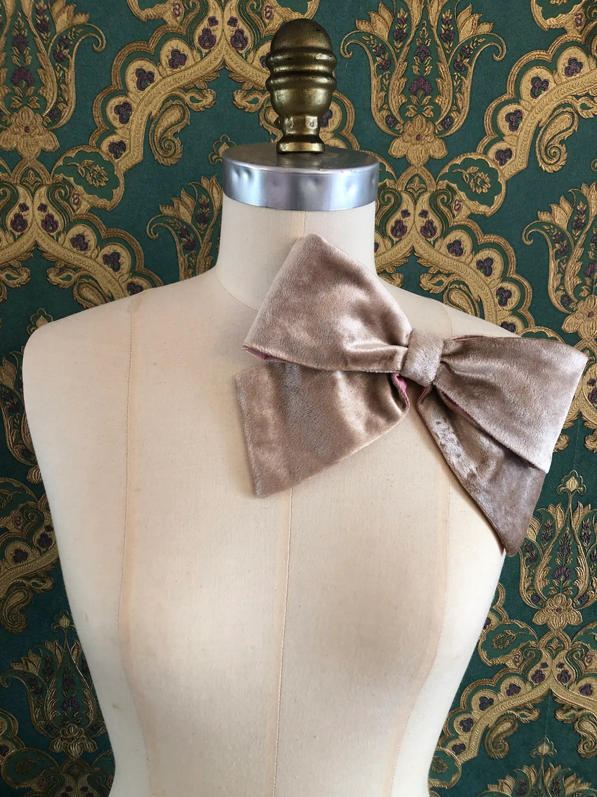 Broochella handmade Large Velvet Bow Brooch in luxurious champagne velvet with contrasting pink lining, styled asymmetrically on a cream mannequin against ornate green and gold wallpaper.