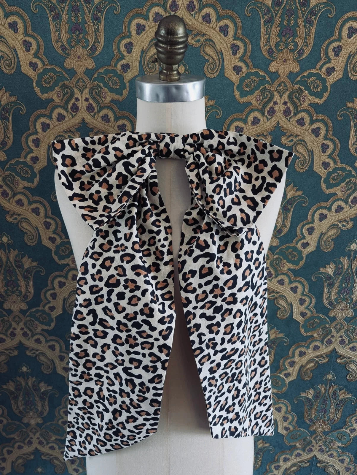 Broochella large, cotton, handmade leopard pint bow brooch. It is pinned to a mannequin. The background is green and gold damask wallpaper.
