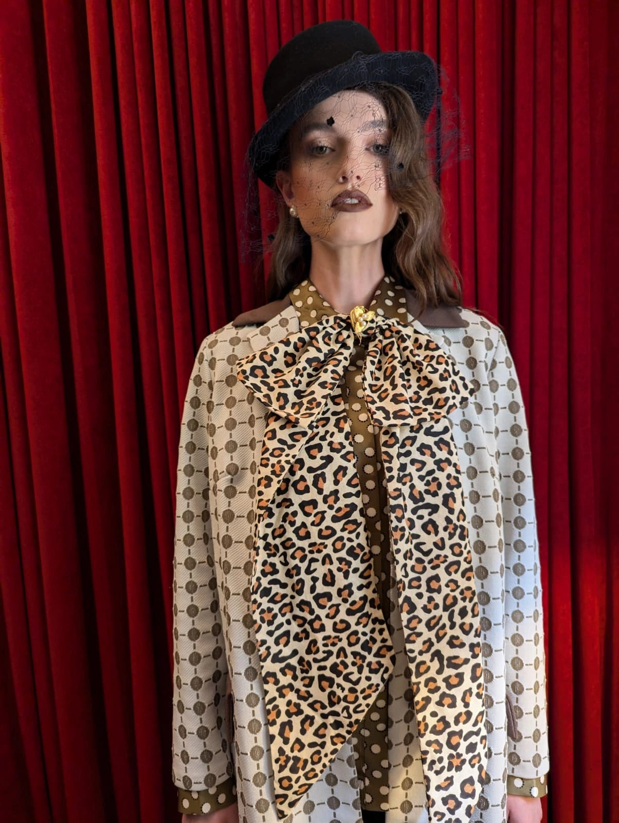 A model wearing a vintage-style beige and brown patterned jacket with a large broochella leopard-print bow tie, a black hat, and a black veil, standing in front of a red curtain.