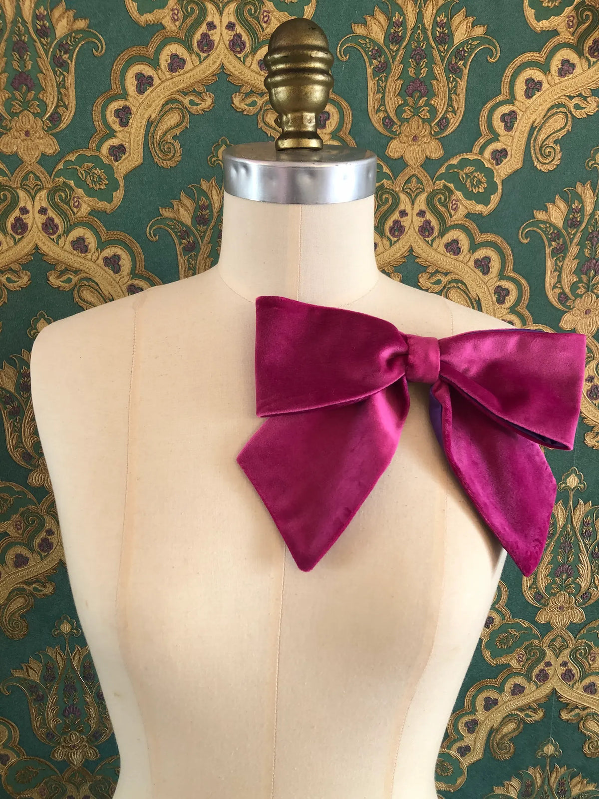 Broochella handmade Large Velvet Bow Brooch in rich magenta velvet with contrasting purple edging, styled asymmetrically on a cream mannequin against ornate green and gold wallpaper.